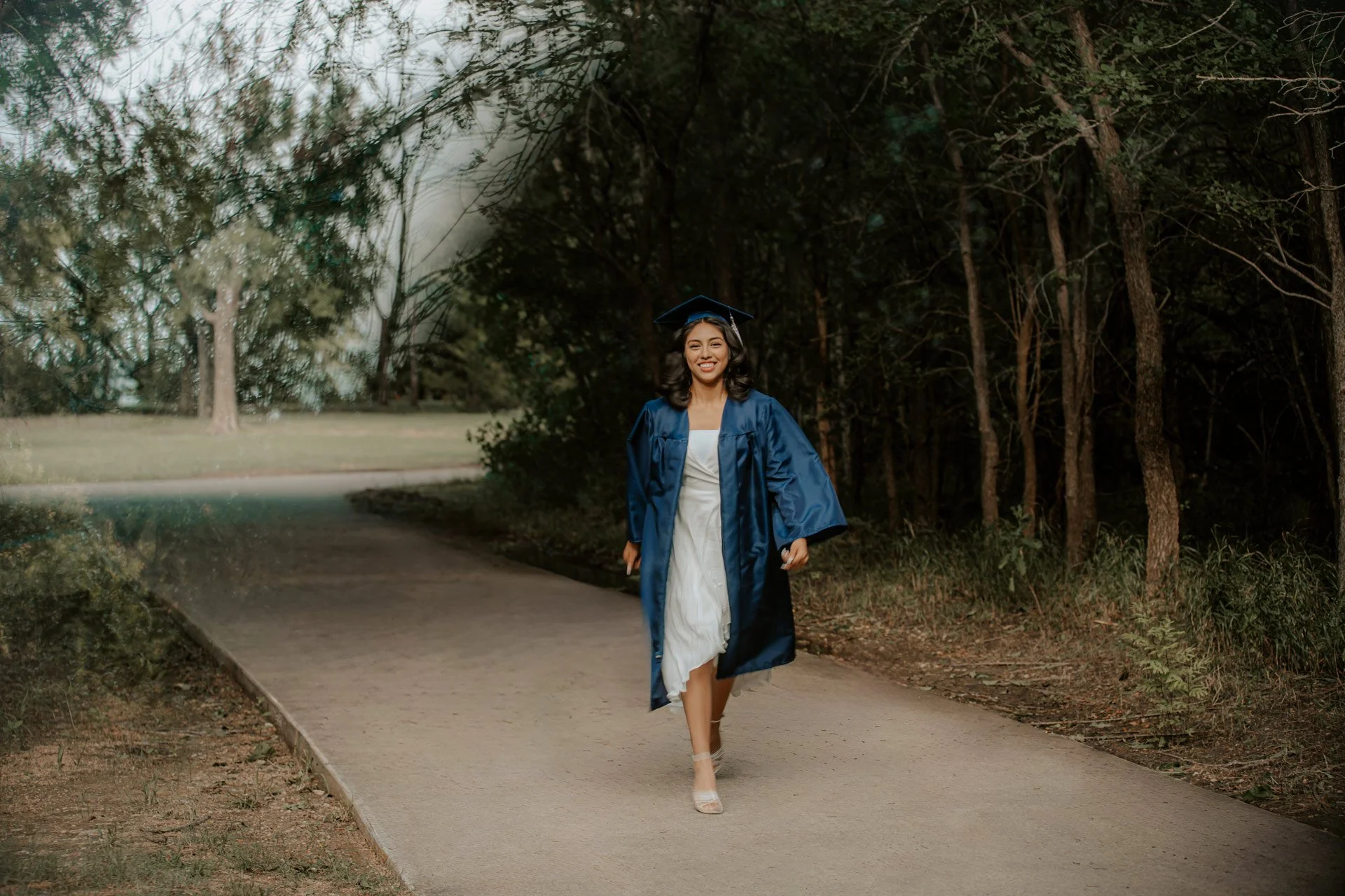A young woman in a white dress and a blue graduation gown and cap is walking on a paved path through a wooded area, smiling at the camera.