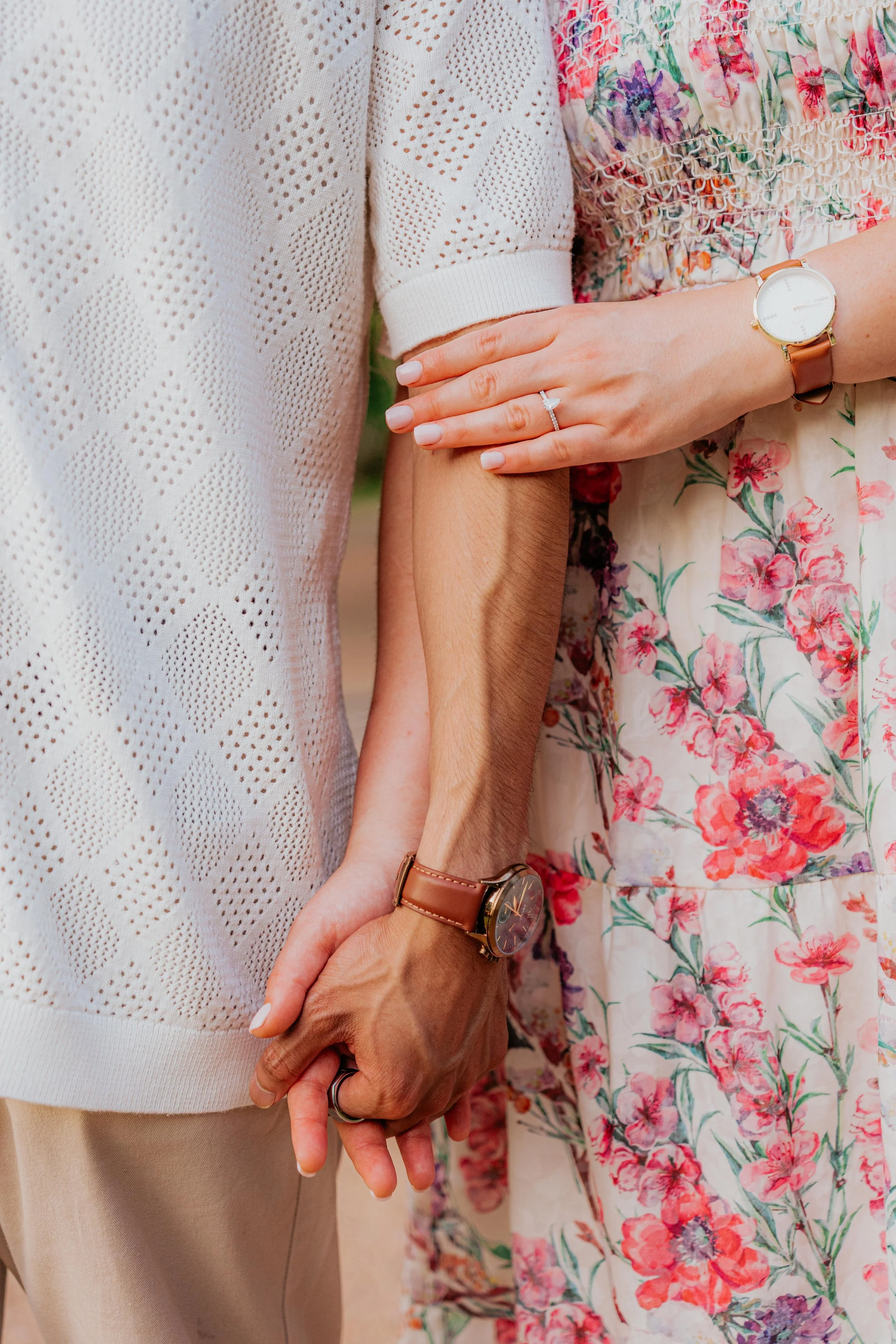 Close-up of a couple holding hands. The woman is wearing a floral dress and a watch, and the man is in a knitted white top and a watch.