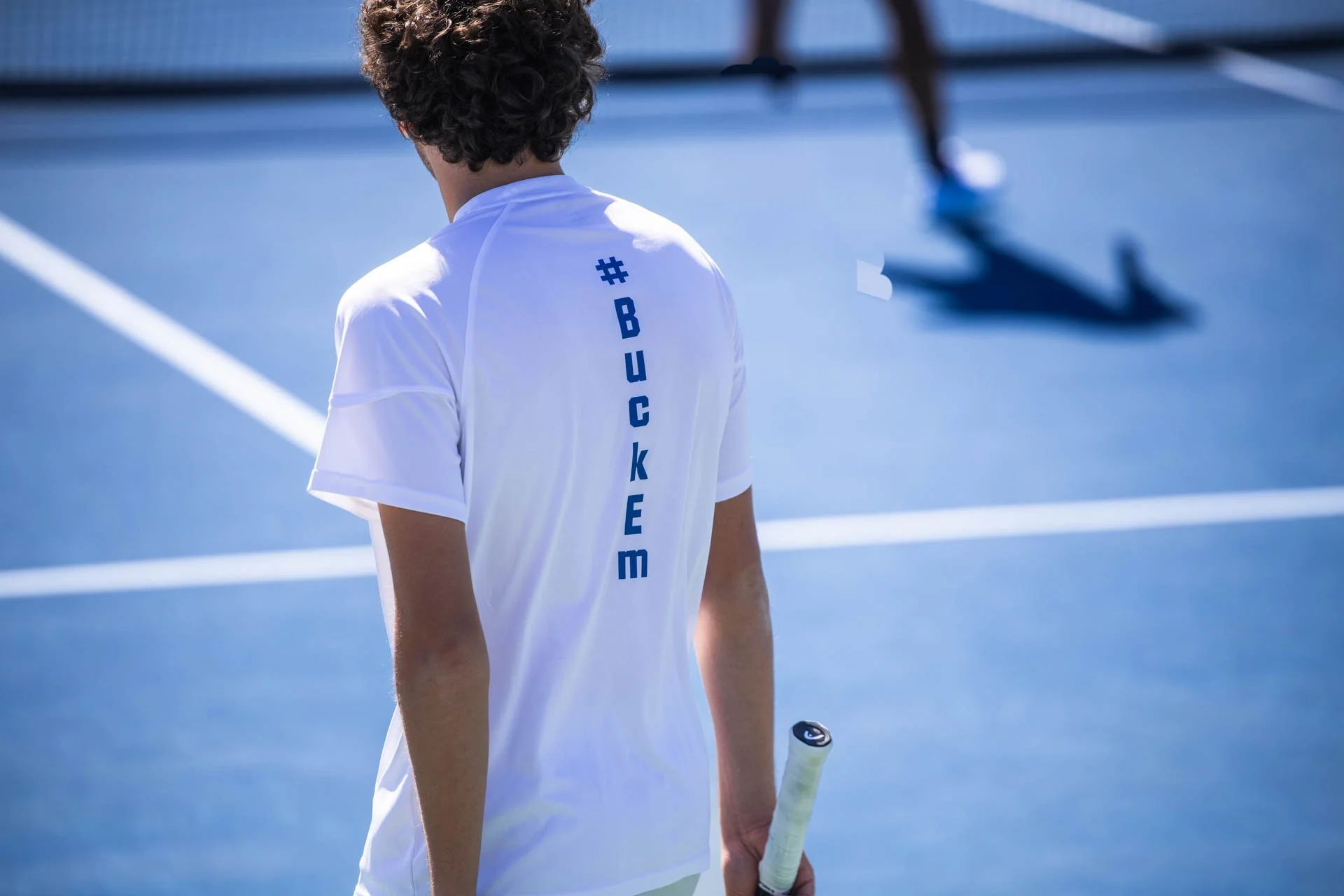 Back view of a young man with curly hair wearing a white sports shirt with '#BUCKEEm' written vertically on the back, holding a tennis racket, standing on a blue tennis court.