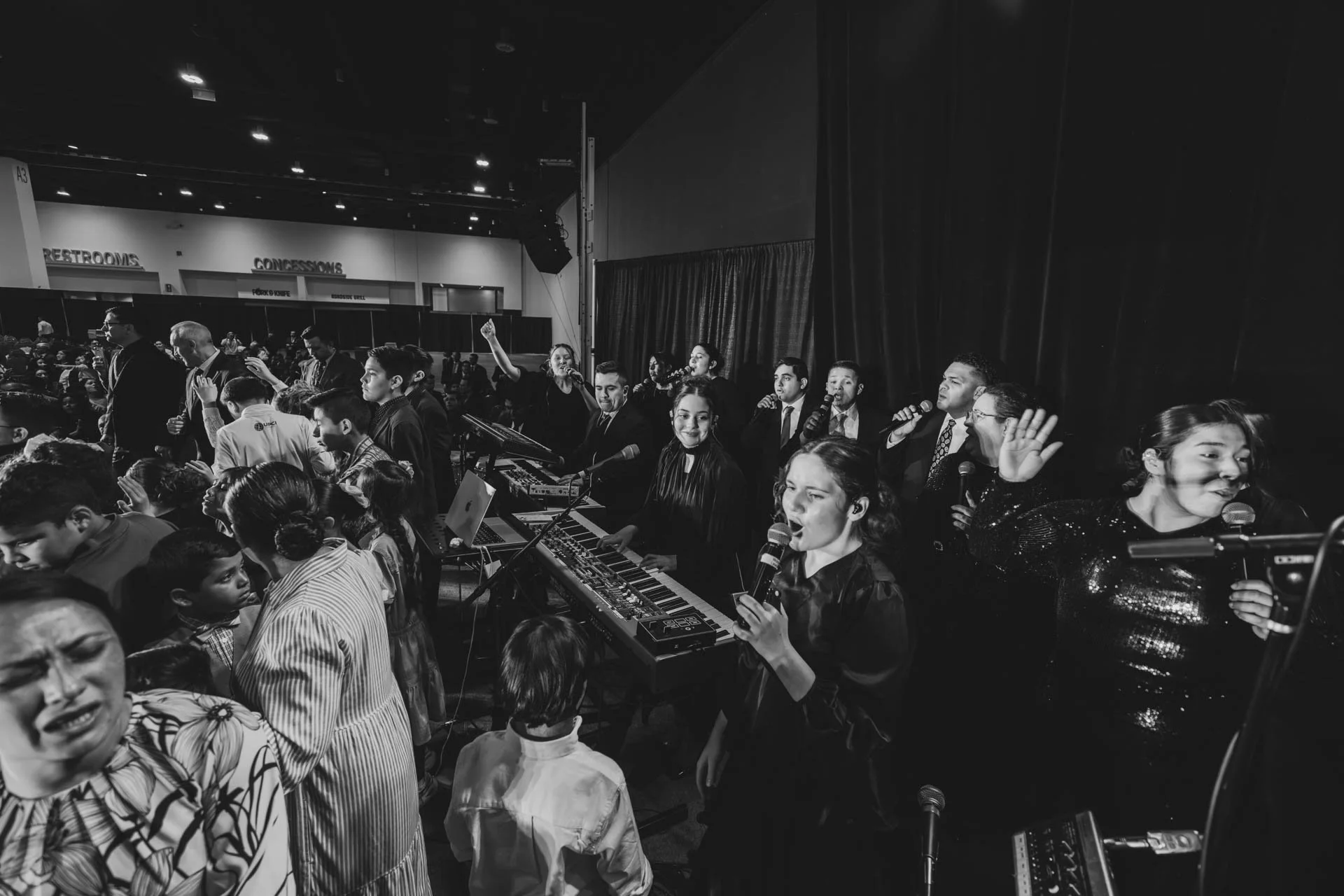 A black-and-white photo of a large group of musicians and singers performing on stage at an event, with an audience and a venue entrance in the background.