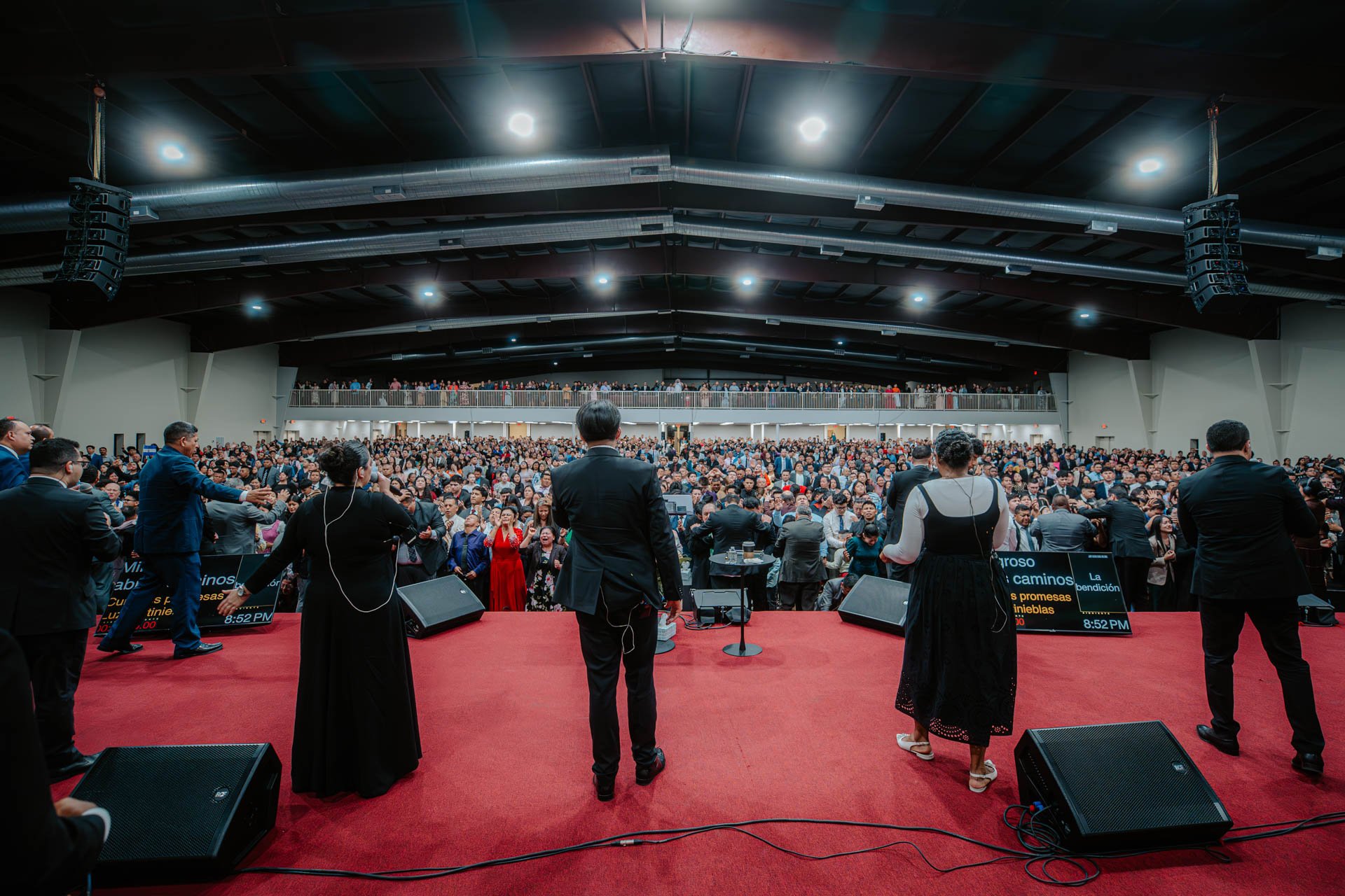 Church service or religious gathering in a large auditorium, with a group of people on stage facing a crowded congregation.