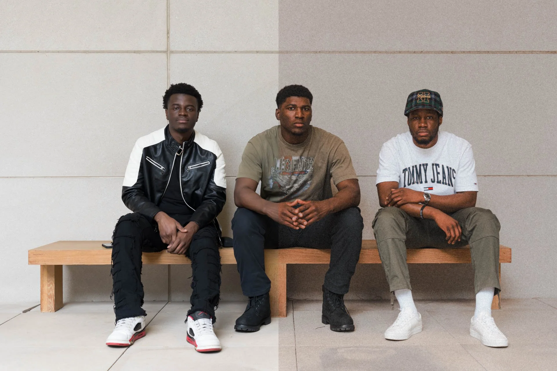 Three young men sitting on a wooden bench against a plain beige wall, dressed in casual streetwear