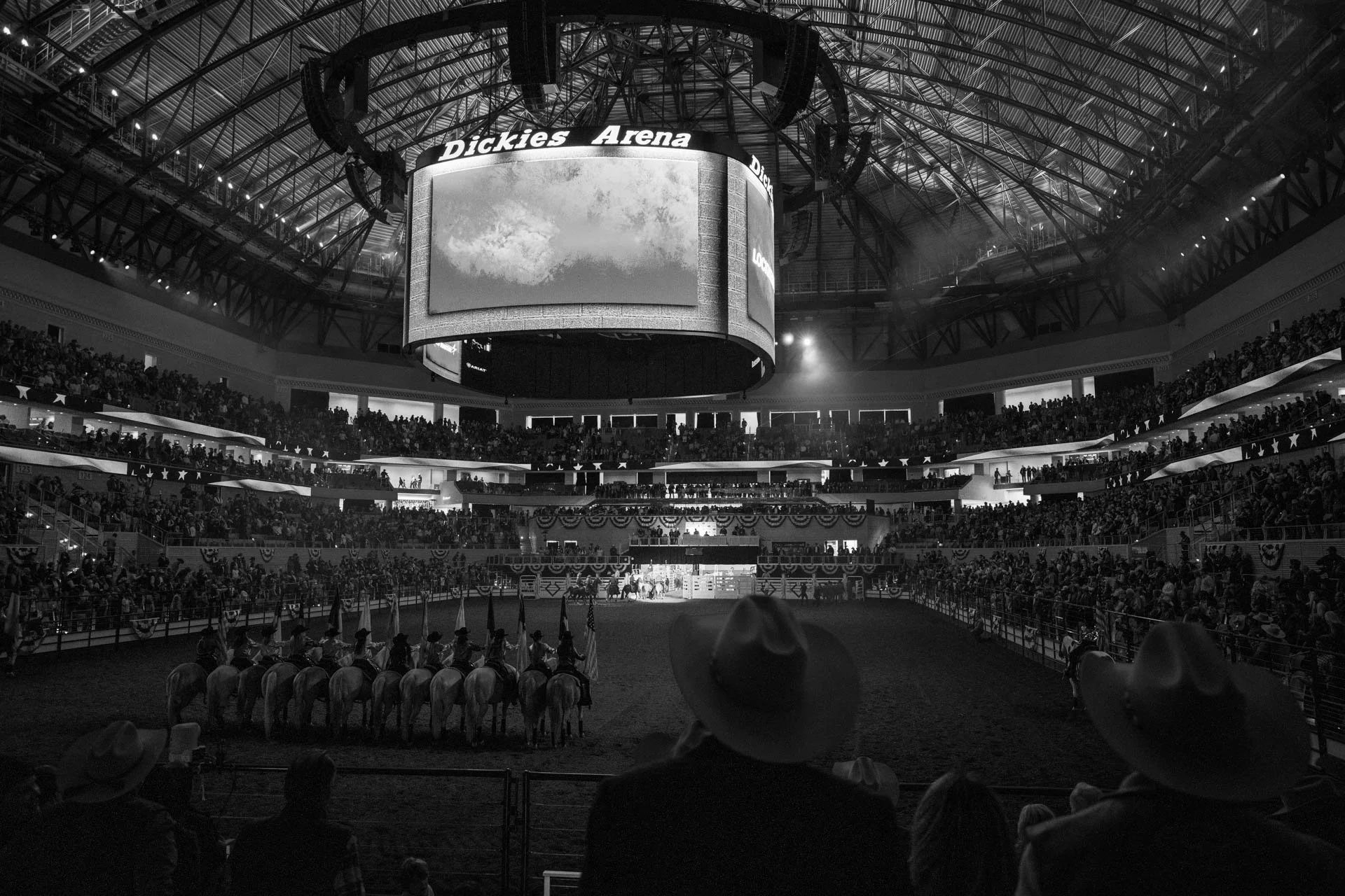 A large indoor arena filled with spectators, with a horse-mounted parade or performance on the arena floor, and a big screen displaying the venue name, Dickies Arena, hanging from the ceiling.