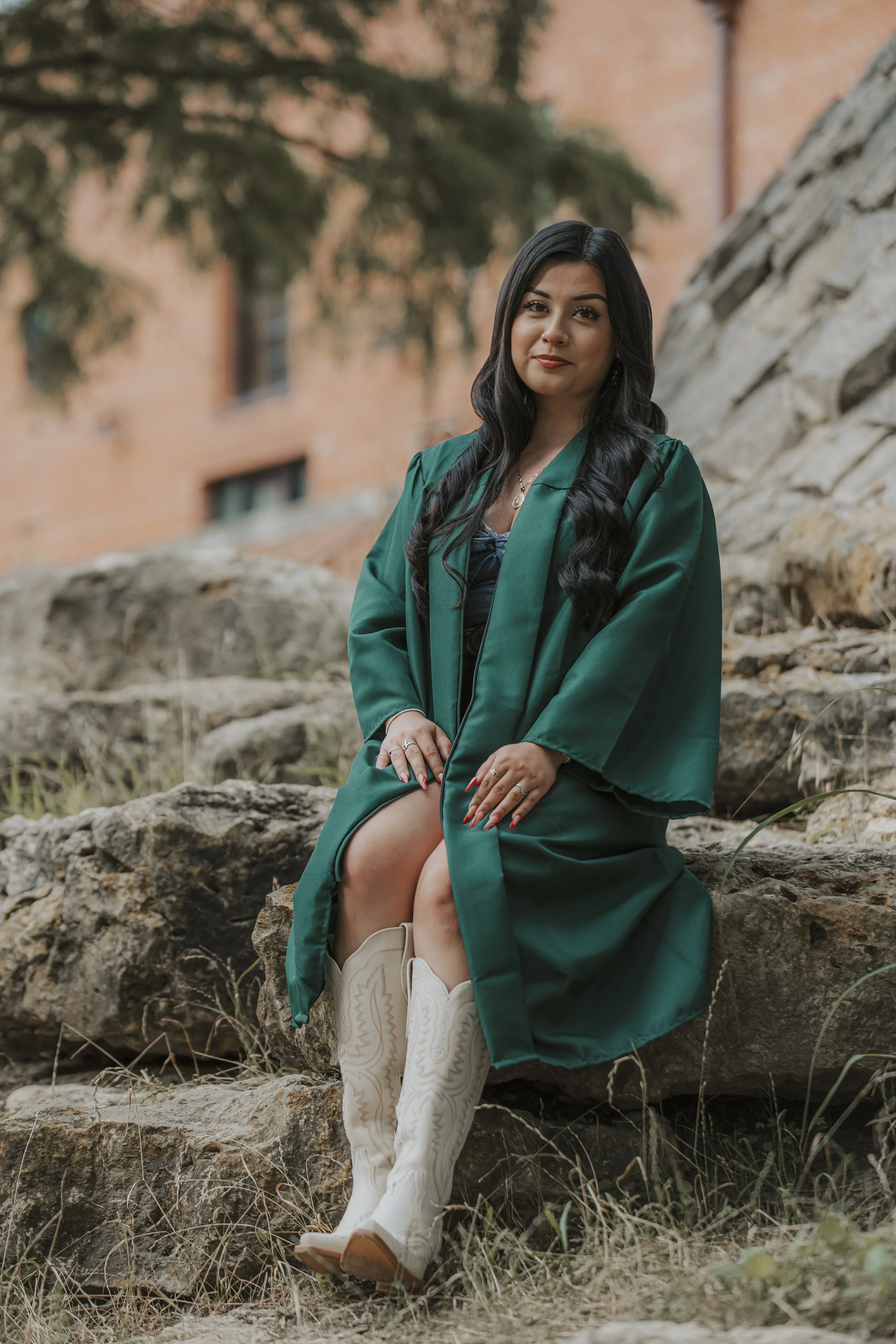 A woman in a green graduation gown sitting on rocks outdoors, wearing white cowboy boots with embroidery, with a blurred brick building and trees in the background.