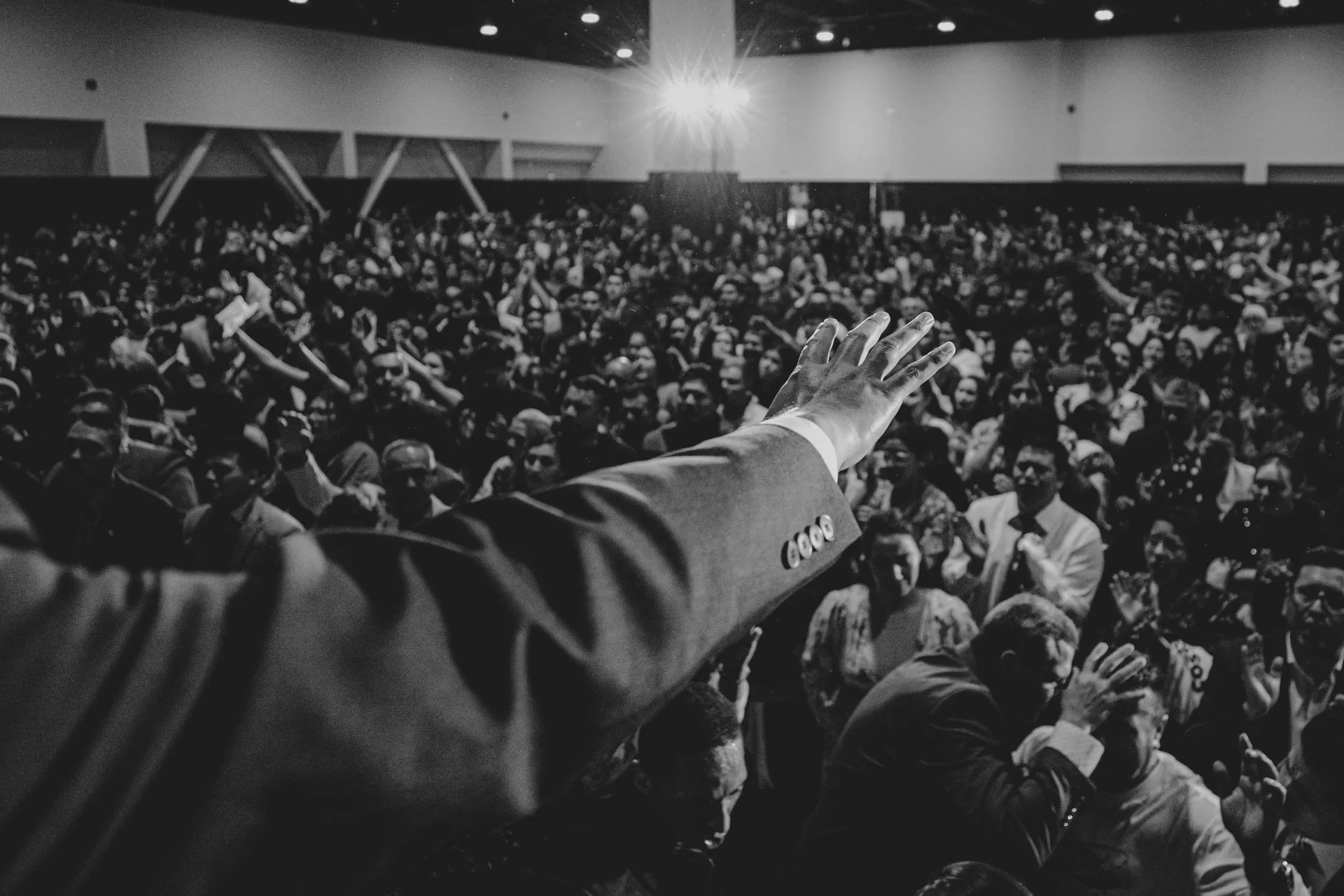 A person in a suit raising their hand towards a large crowd in an auditorium, black and white photo.