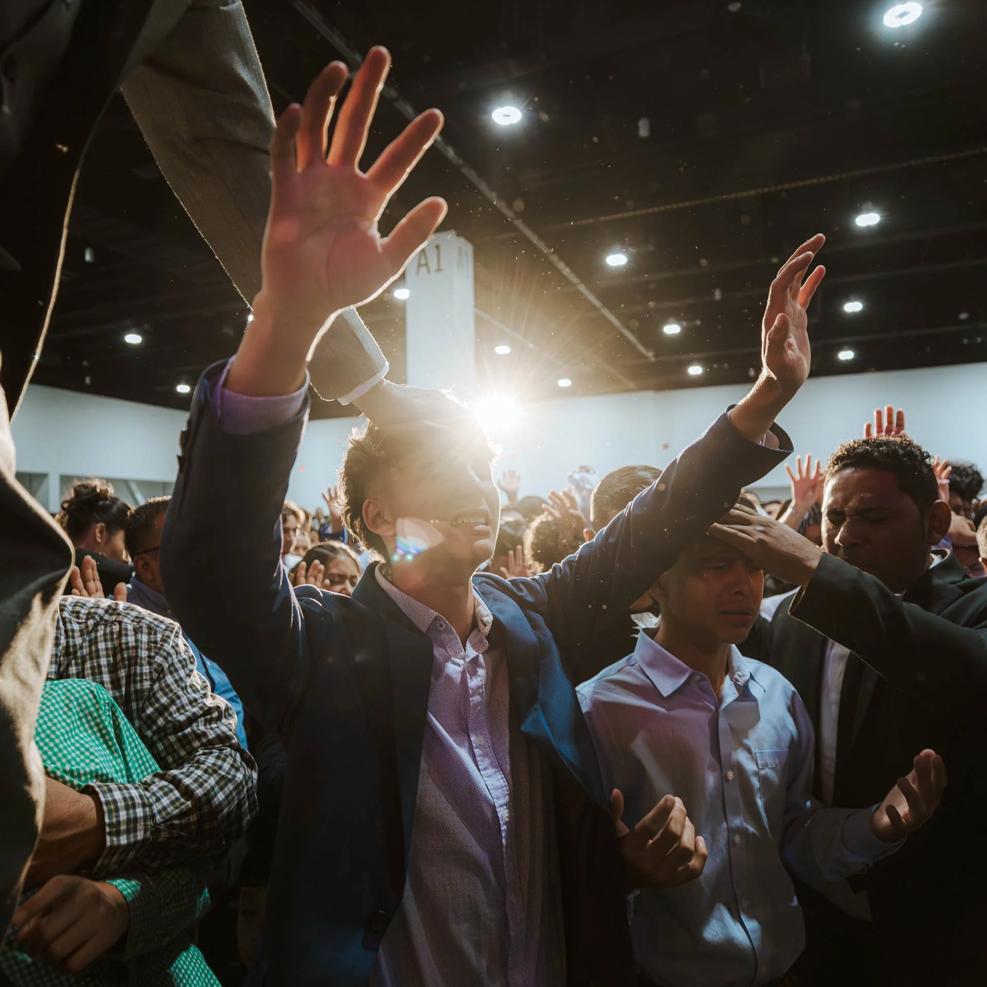 A person with short hair in a blazer and button-up shirt is being prayed over by two men at a crowded indoor event. The person is raising their hands in the air and smiling, with a bright light behind them.