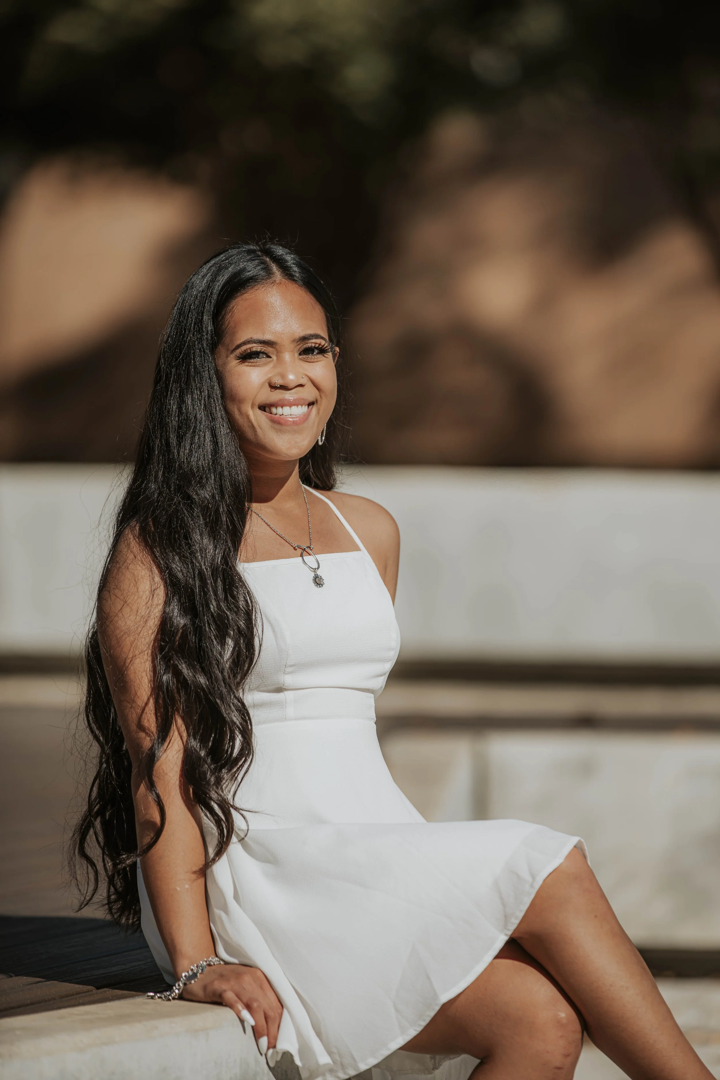Young woman with long dark hair sitting outdoors on a wooden bench, smiling at the camera, wearing a white dress and jewelry.