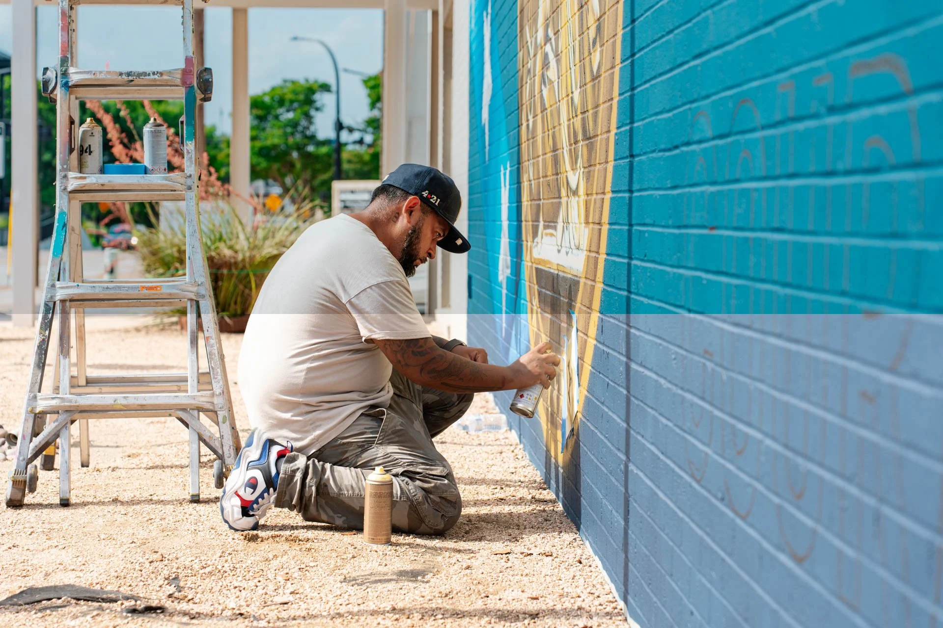 Man spray painting a colorful mural on a brick wall outdoors with a ladder nearby.