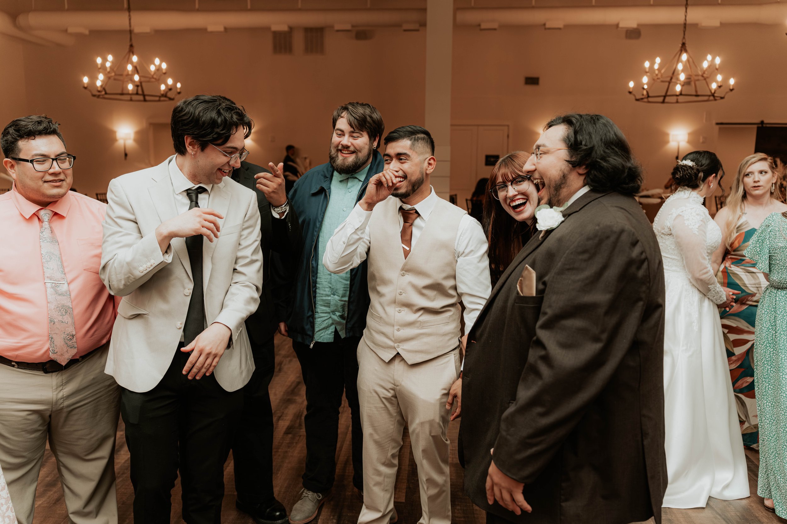 Group of people at a celebration, dressed in formal and semi-formal attire, laughing and enjoying a moment together in a warmly lit room with chandeliers.