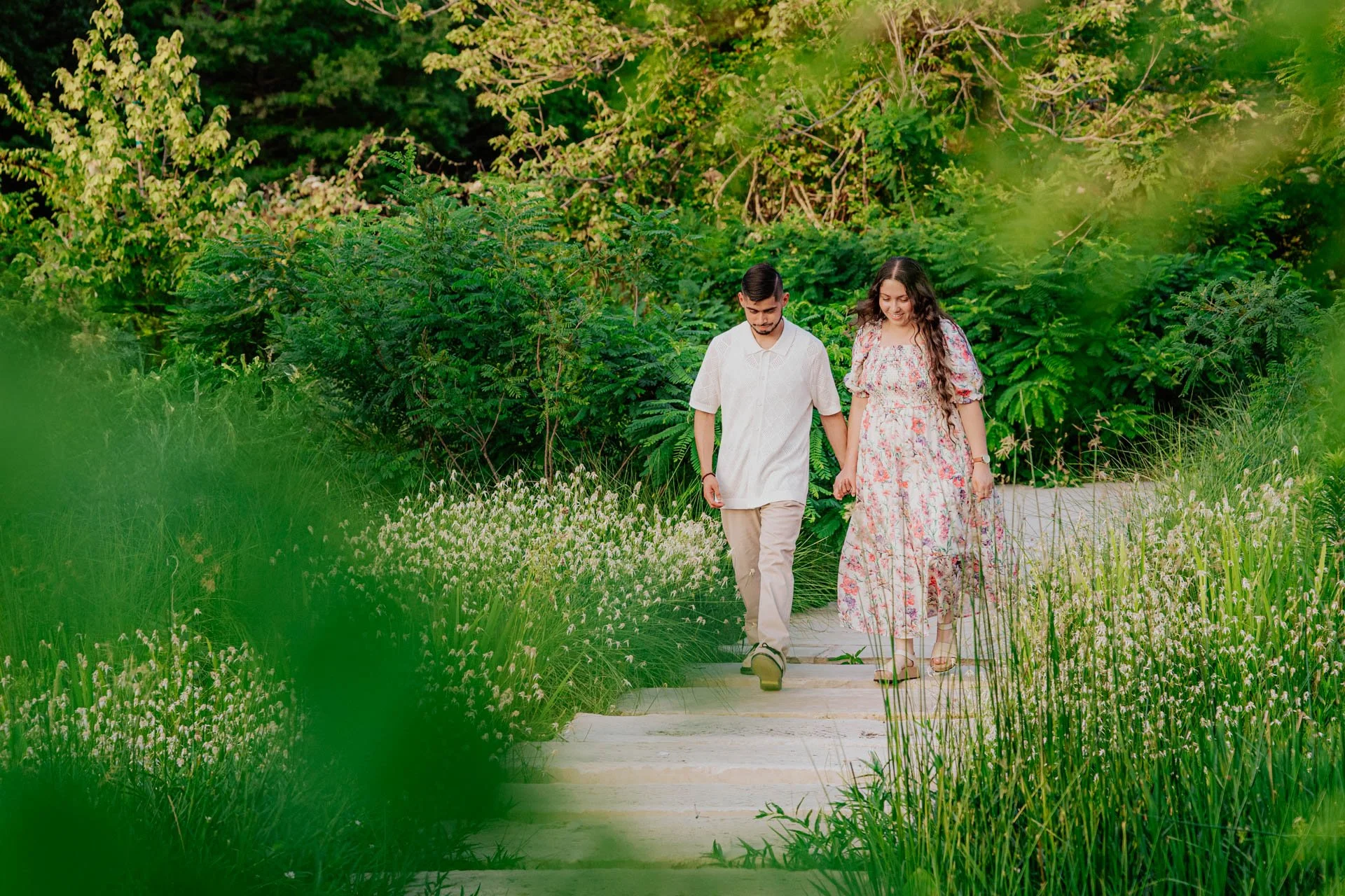 A young couple holding hands and walking along a garden path surrounded by green plants and flowers.