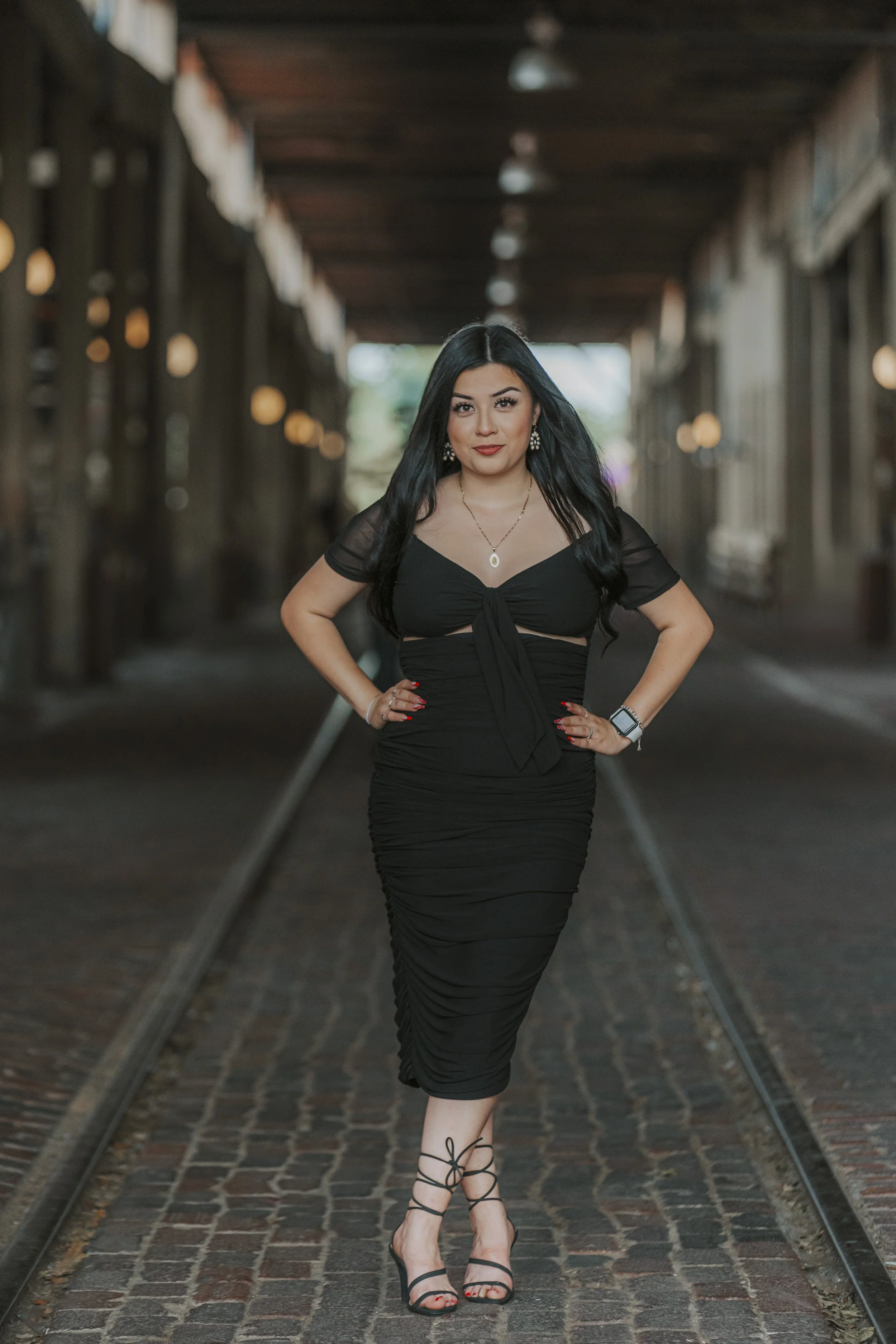 A woman with long black hair wearing a black dress and black strappy high heels stands on a cobblestone street with train tracks, posing with hands on hips and looking at the camera.