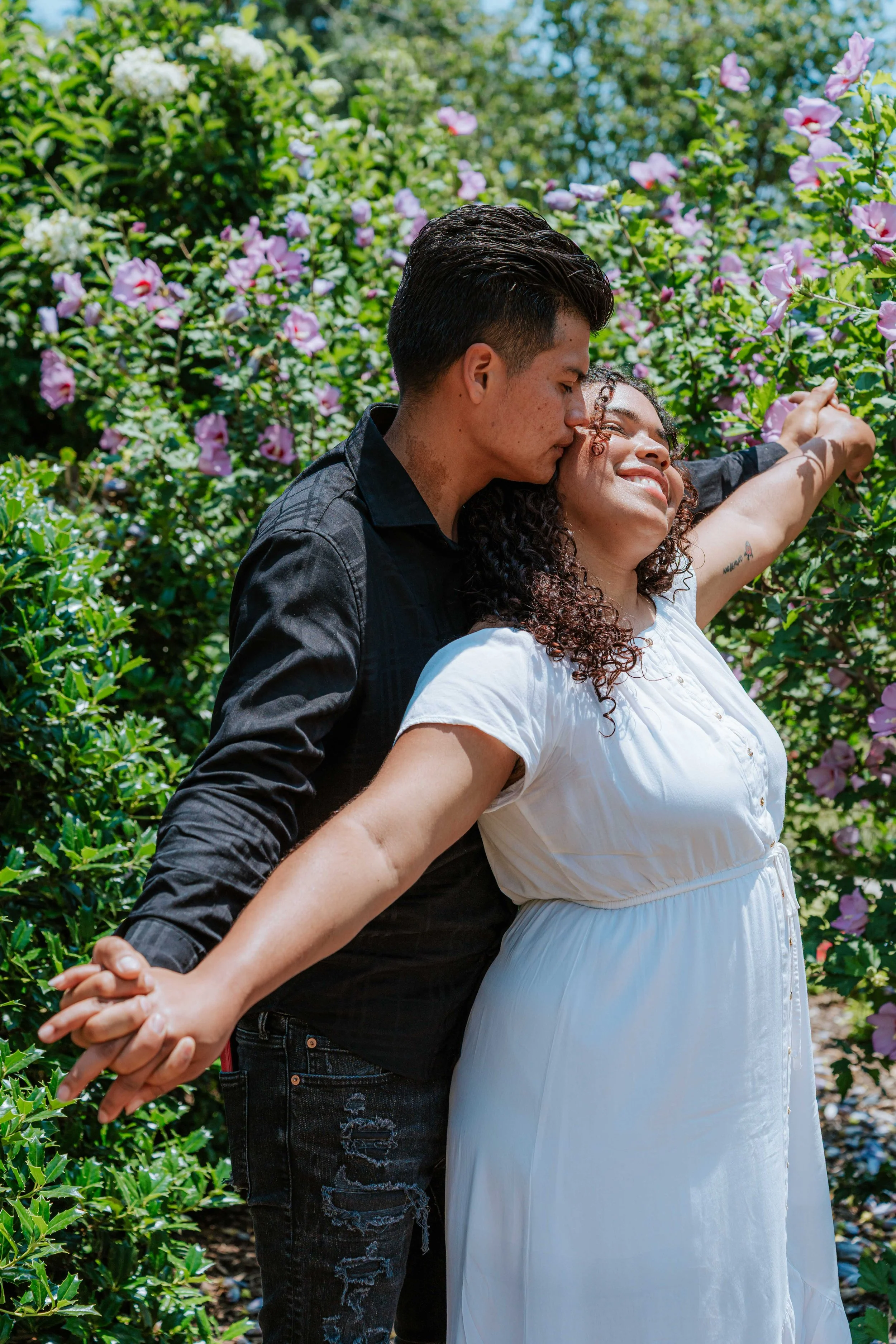 A couple is standing in a garden among blooming pink and white flowers, holding hands and embracing. The man is kissing the woman on the forehead, and both are smiling, with the woman showing her profile and the man facing away from the camera.