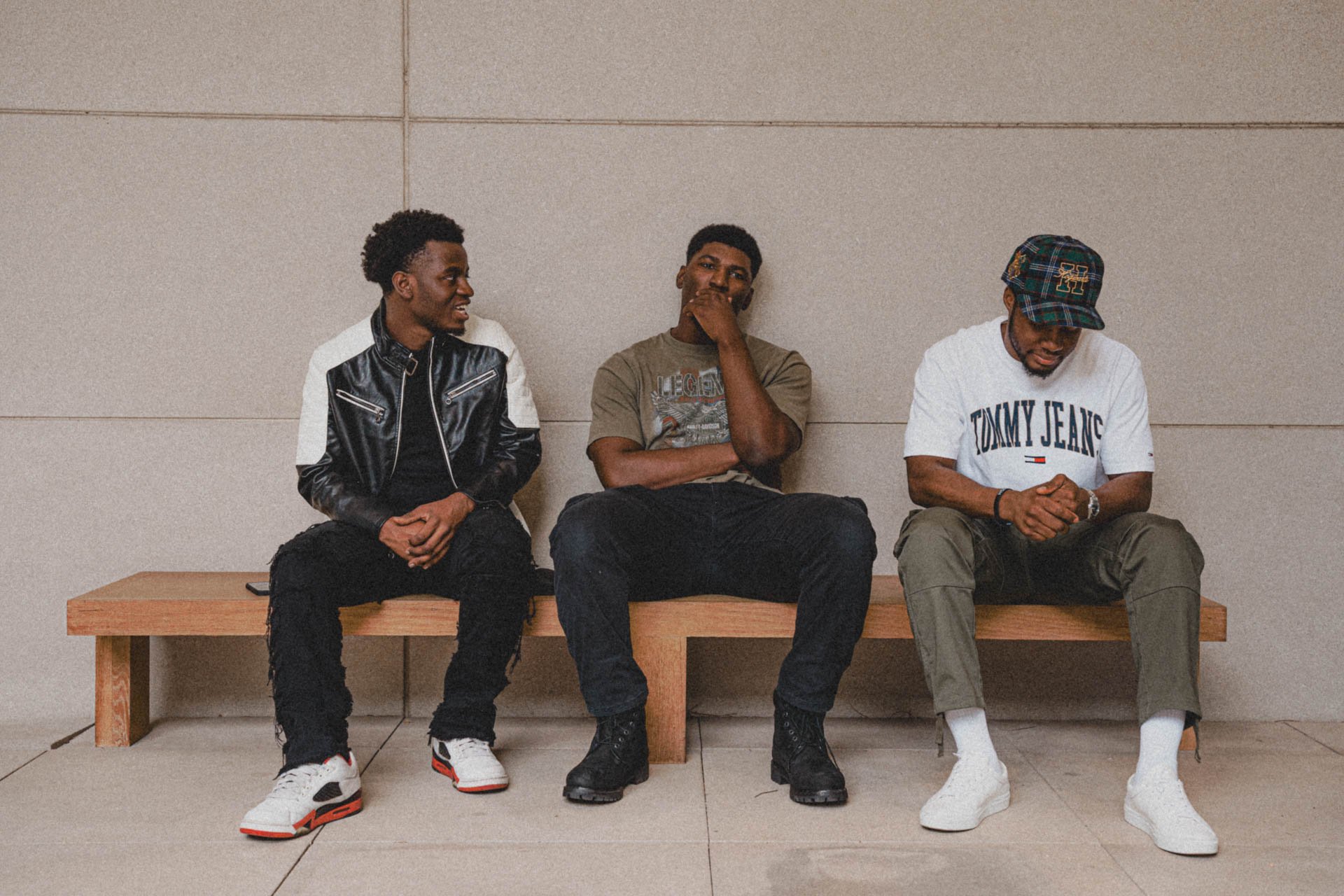 Three men sitting on a wooden bench against a beige wall. The man on the left is wearing a black and white jacket with white sneakers. The man in the middle is wearing a gray T-shirt and black pants, with his hand covering his mouth. The man on the r
