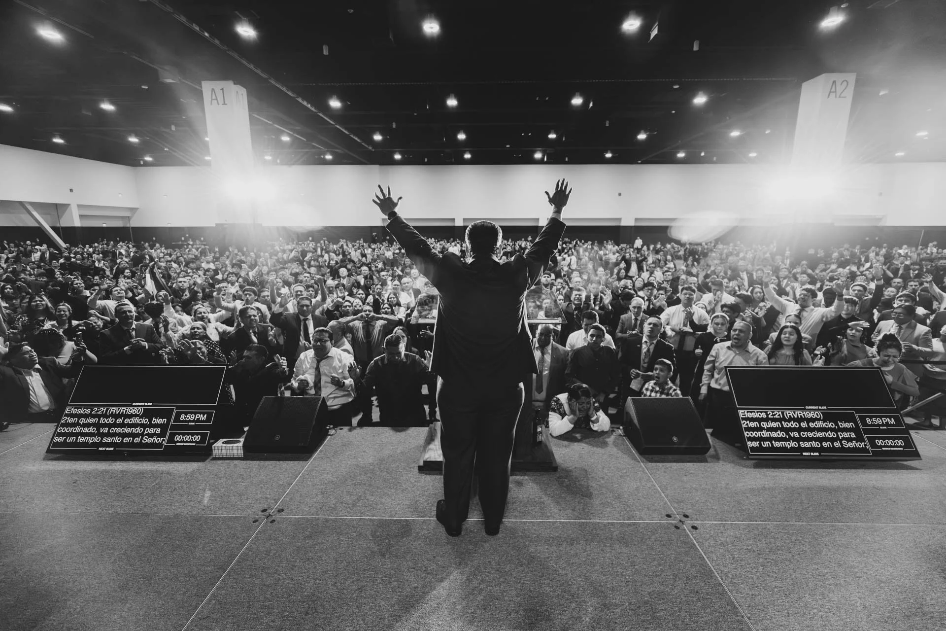 A man on stage with arms raised, addressing a large crowd in an indoor auditorium. The audience appears engaged, and there are two screens displaying text and a clock.