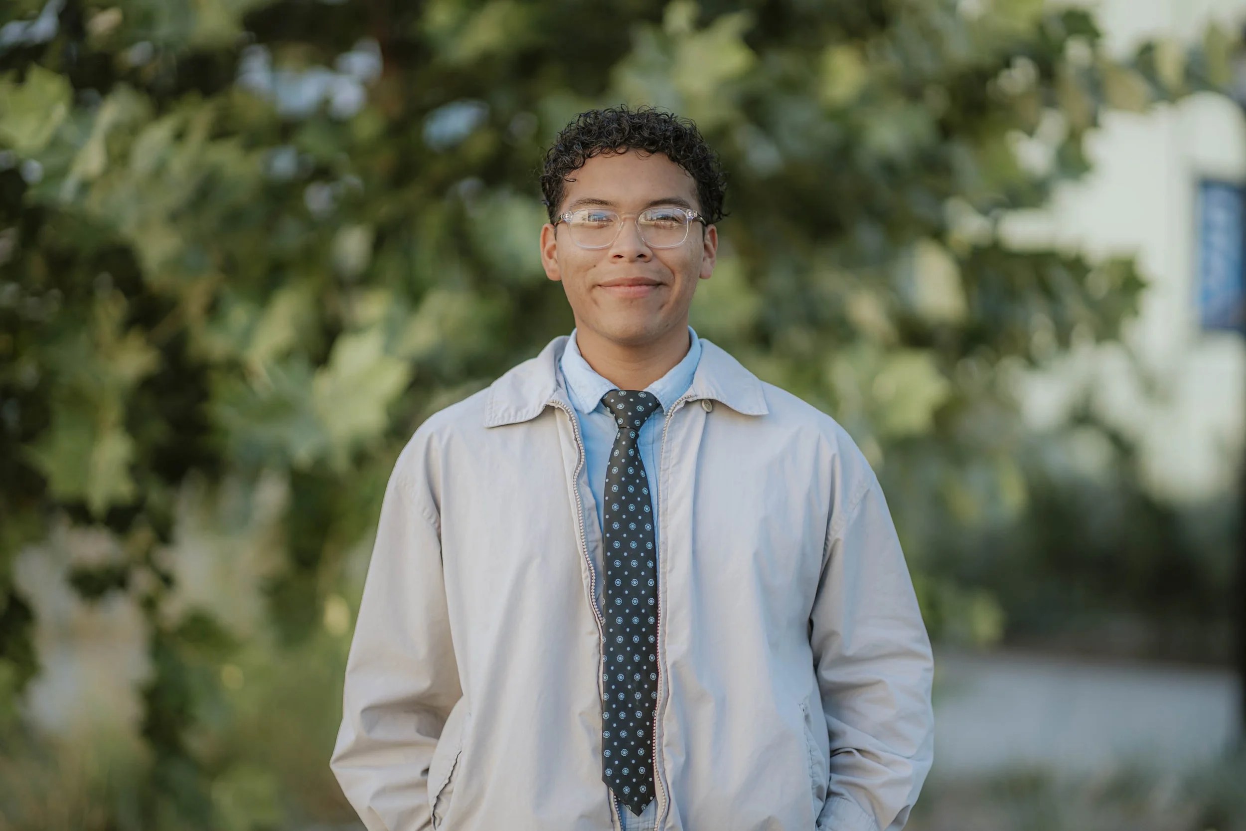 A young man with curly hair and glasses standing outdoors in front of green trees, wearing a light jacket, a blue shirt, and a polka dot tie.
