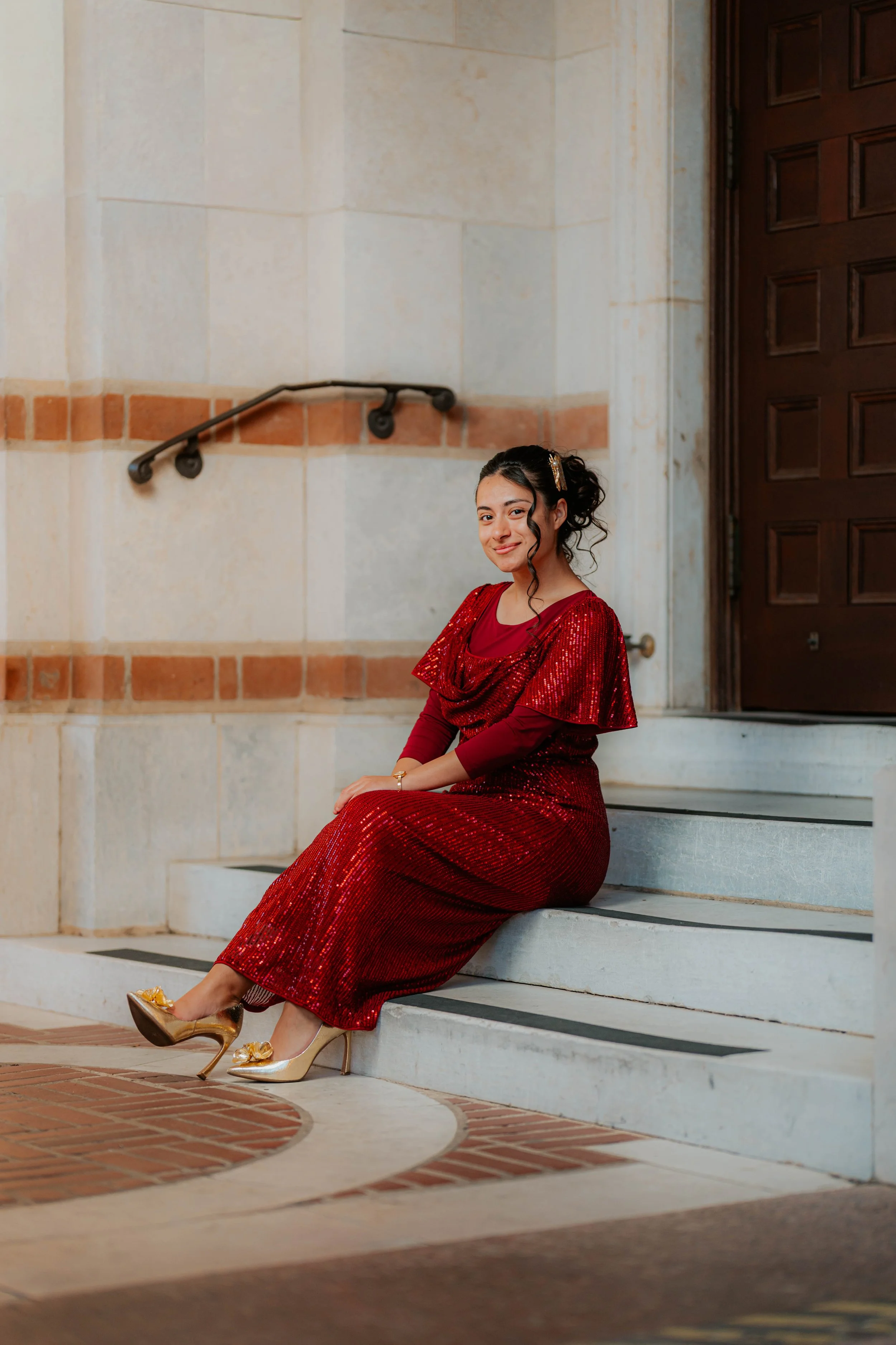 A young woman wearing a red, sparkly dress and gold high heels sits on white stone steps outside a building with a wooden door, smiling at the camera.