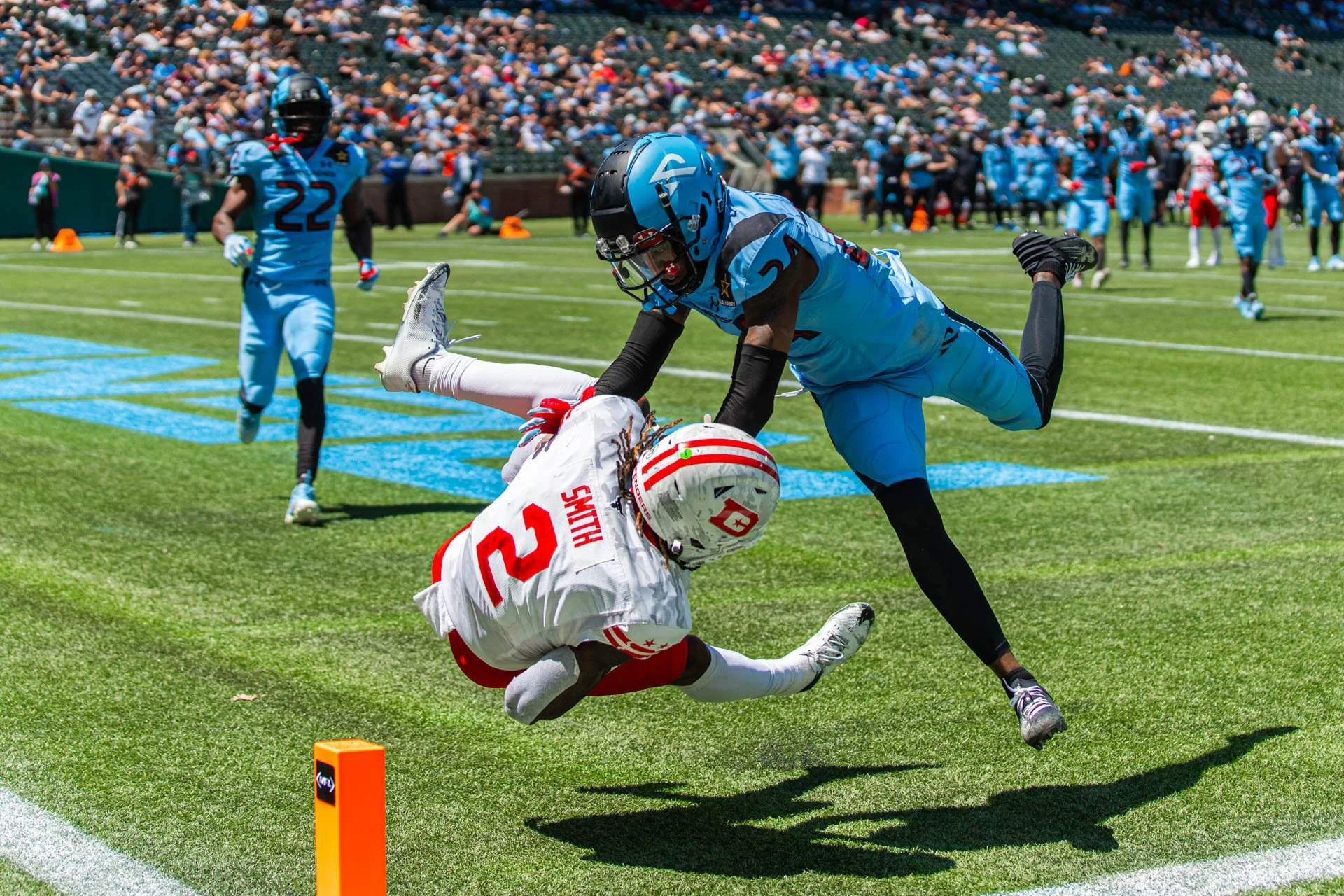 Football player in white jersey tackled by player in blue jersey during game on a green field with crowd in the stands.
