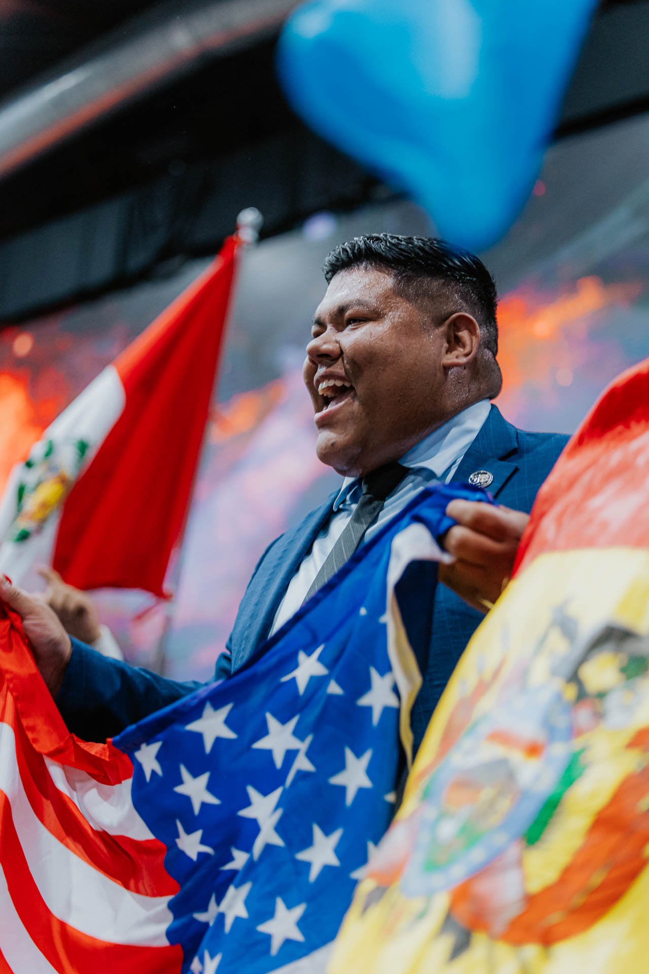 A man in a suit holding up flags, smiling, with patriotic flags including the United States flag, in a celebratory setting.