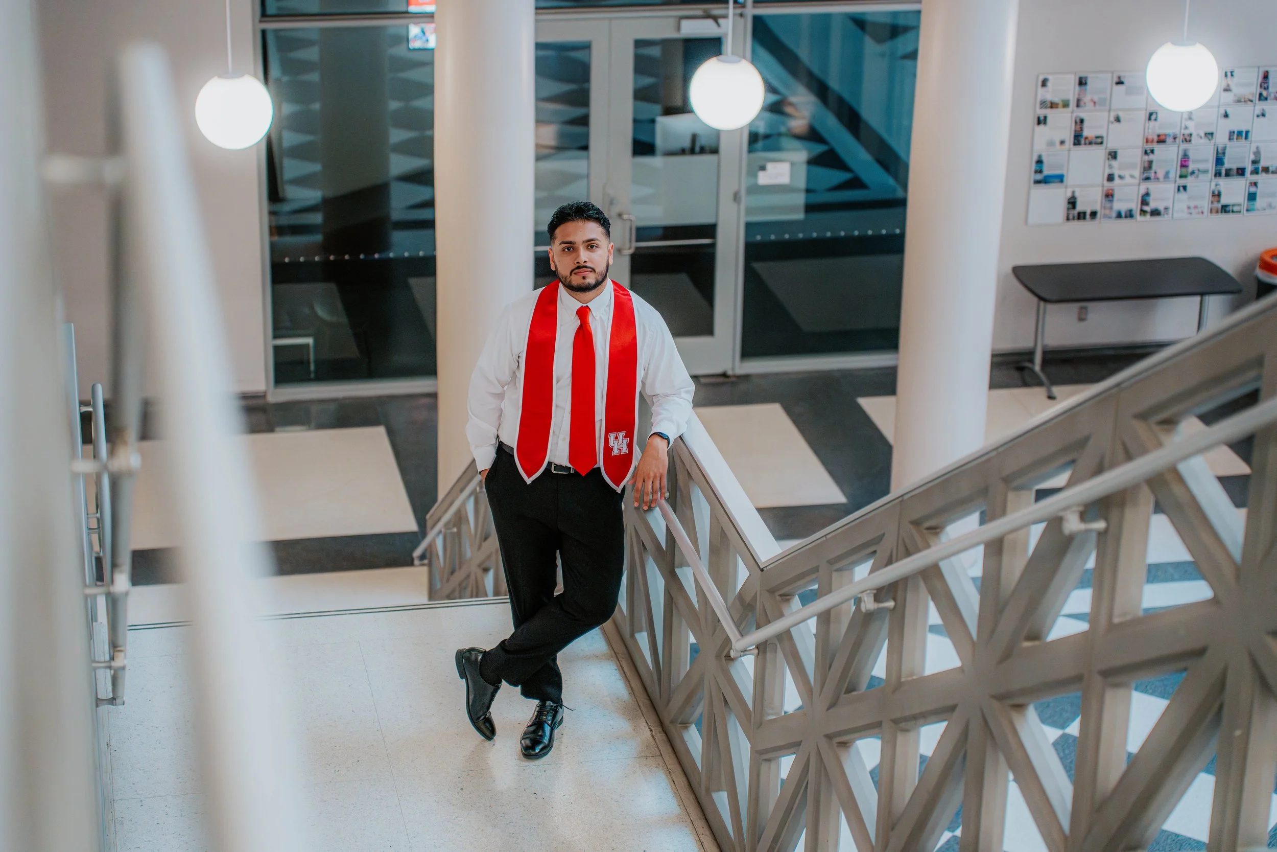 A man in formal attire, wearing a white shirt, red tie, and red scarf with a university logo, stands on a staircase in a modern building, looking at the camera.