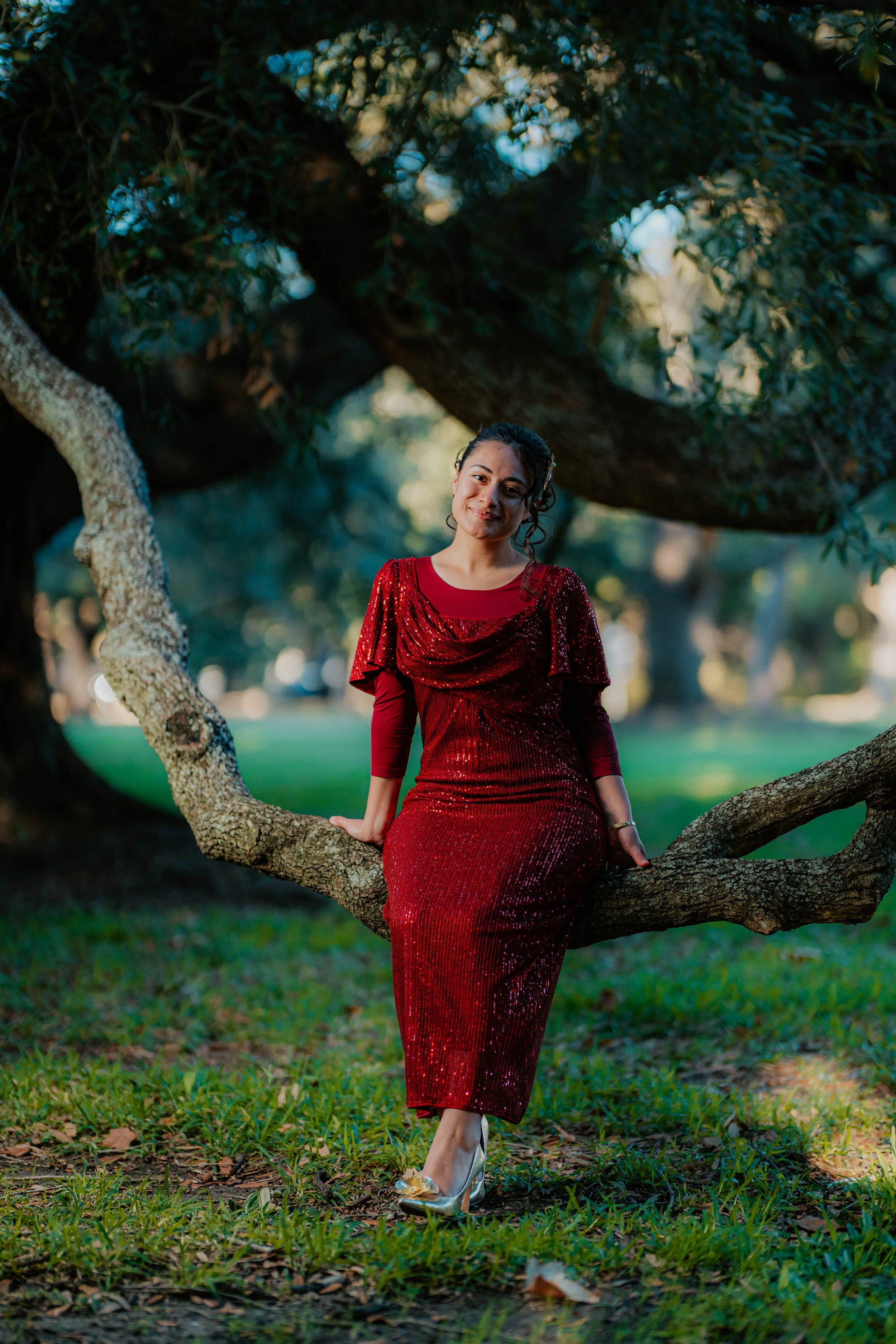 A woman in a red sequined dress sitting on a tree branch outdoors in a park with lush green grass and trees in the background.