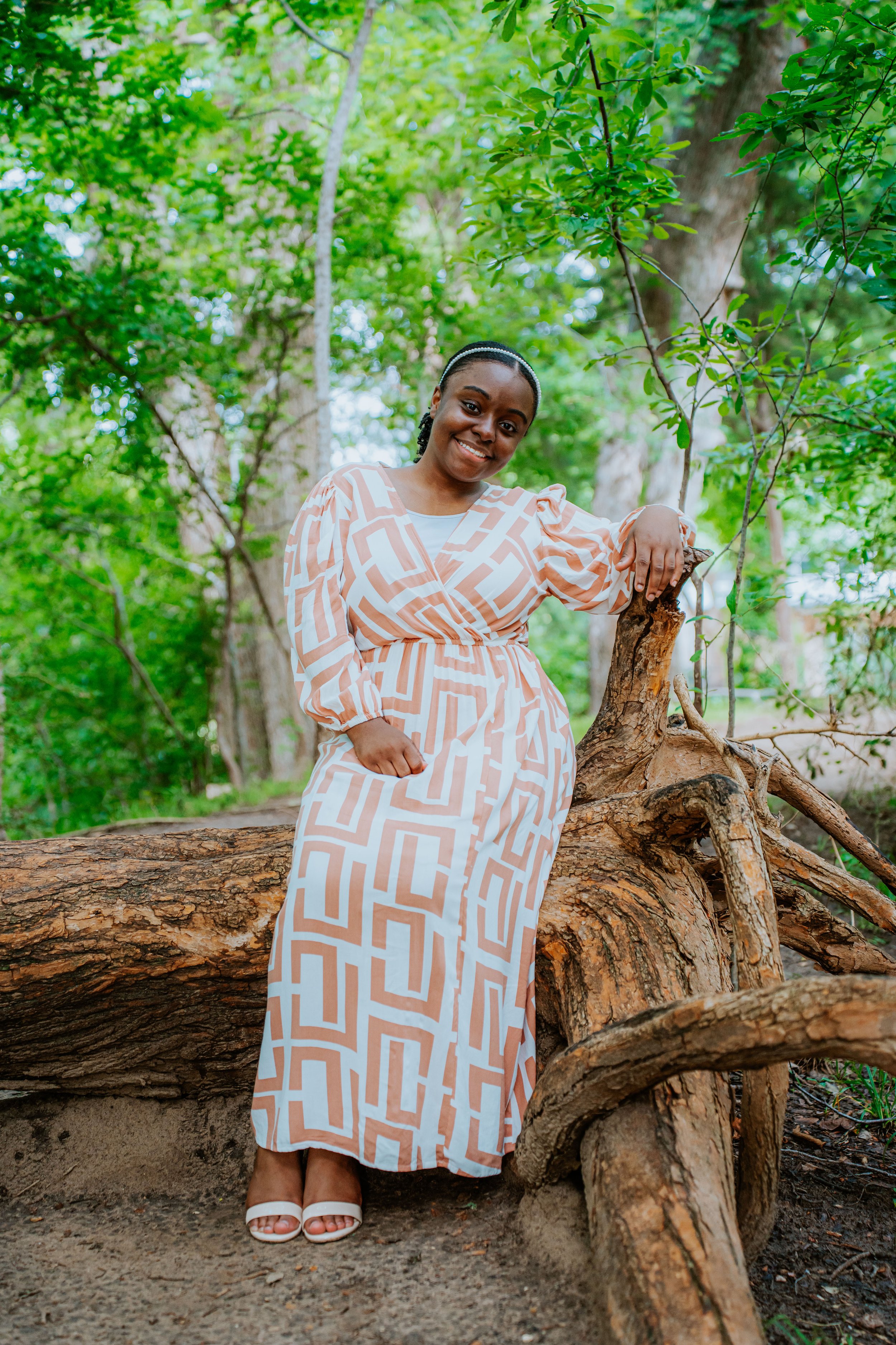 A smiling woman in a patterned dress stands outdoors, leaning on a fallen tree surrounded by lush green trees.