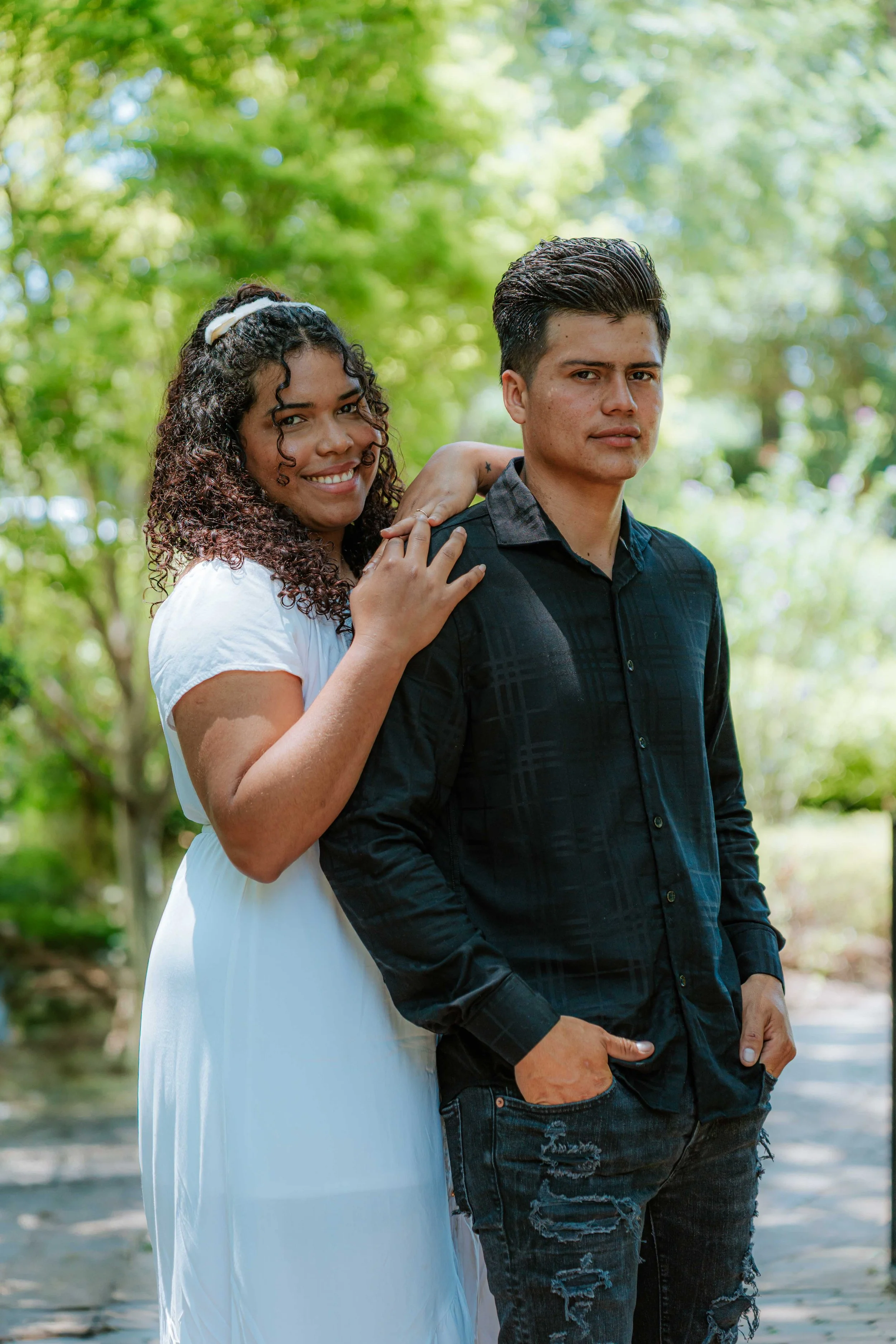 A young couple standing outdoors surrounded by green trees. The woman, with curly hair, is smiling and resting her hand on the man's shoulder. The man has short dark hair, a serious expression, and has his hands in his pockets.
