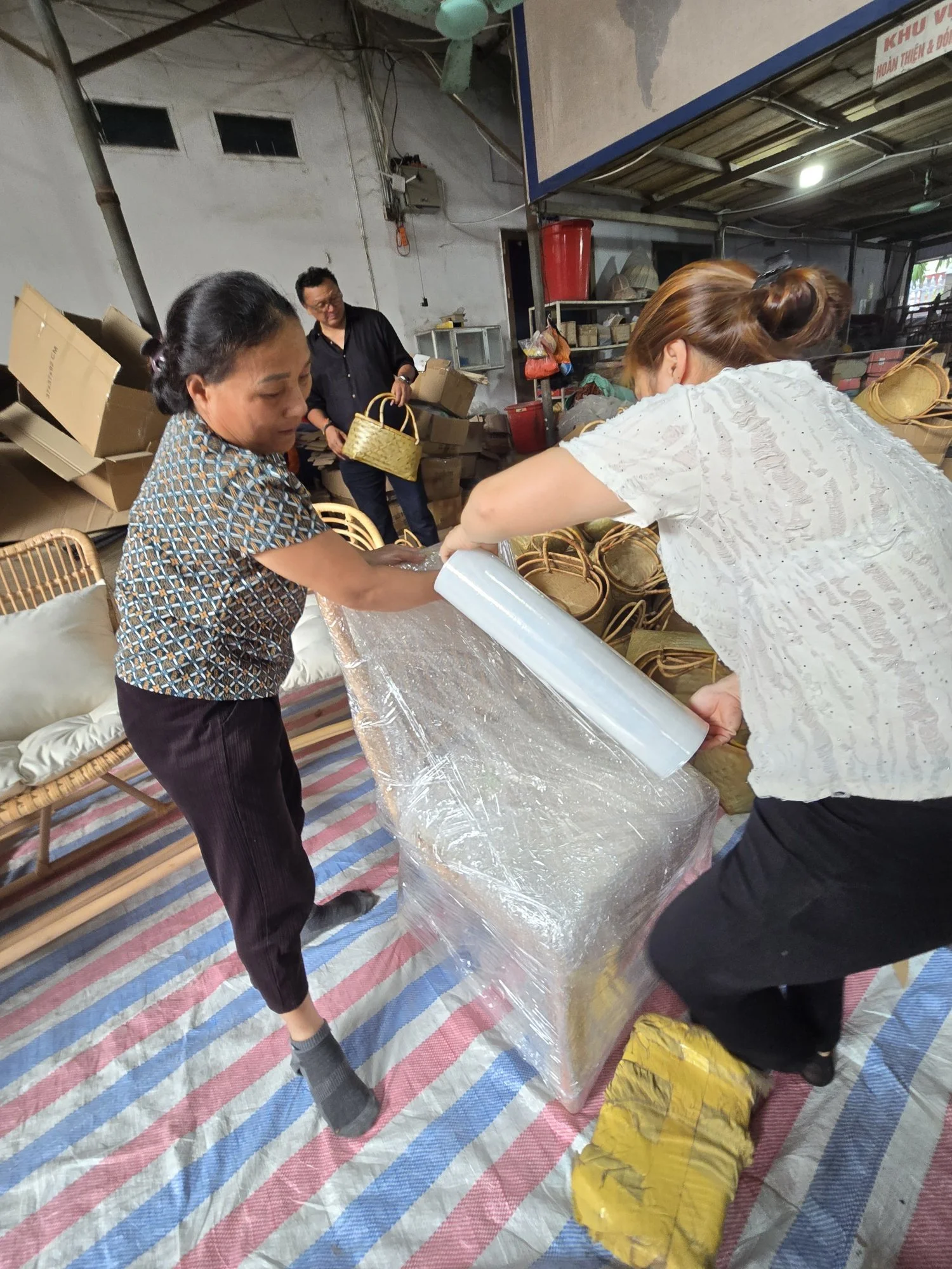 Two women are wrapping a wicker basket in plastic wrap at a workshop or warehouse filled with baskets and cardboard boxes. A man is standing in the background holding a basket and looking at the scene.