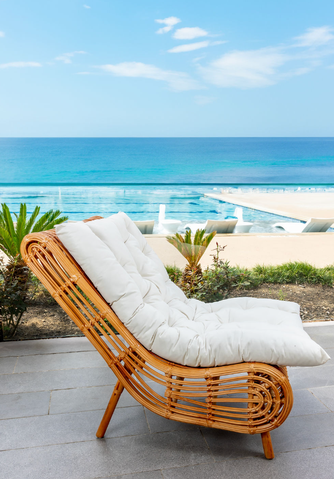 A rattan lounge chair with a white cushion on an outdoor patio overlooking the ocean, with beach chairs by the shoreline and blue sky.