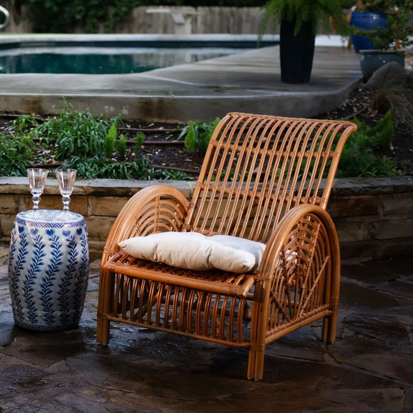 A rattan armchair with a white pillow on it, positioned on a stone patio. Next to the chair is a ceramic side table with a blue leaf pattern, holding two glasses. In the background, there is a garden with green plants and a swimming pool.