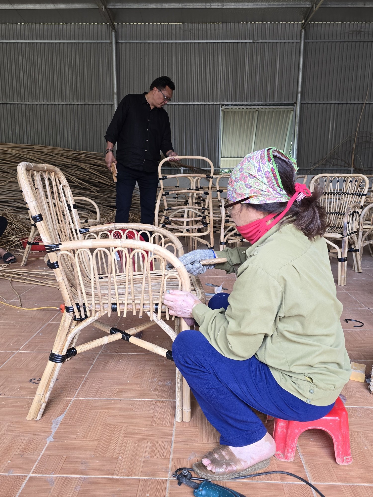 Two people assembling bamboo chairs in a workshop. One person is sitting on a small red stool, working on a chair, while the other stands in the background, arranging more chairs. The workshop has a metal corrugated roof and walls, and wooden materials are stacked against the wall.