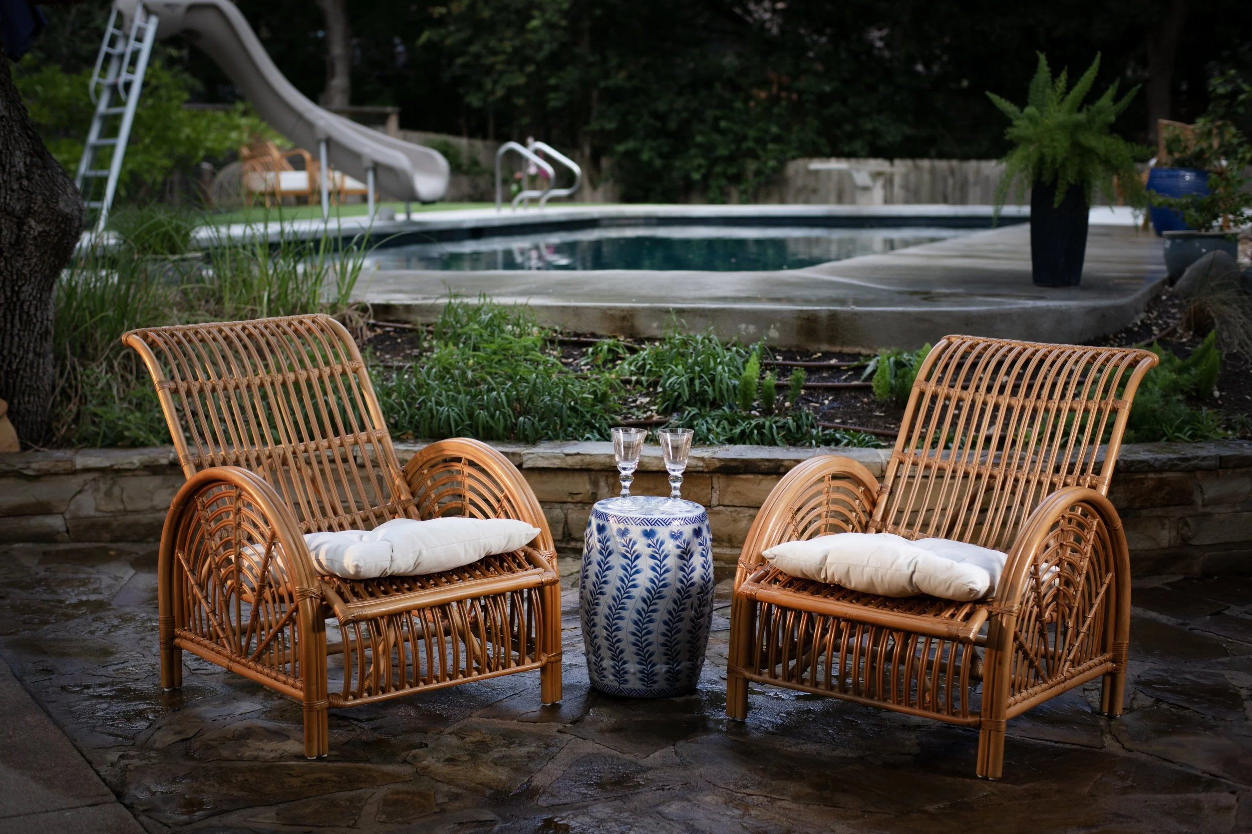 Two rattan chairs with white cushions facing each other with a small blue and white ceramic table in between. On the table are two empty wine glasses. In the background, there is a swimming pool, a slide, a lush green garden, and potted plants.