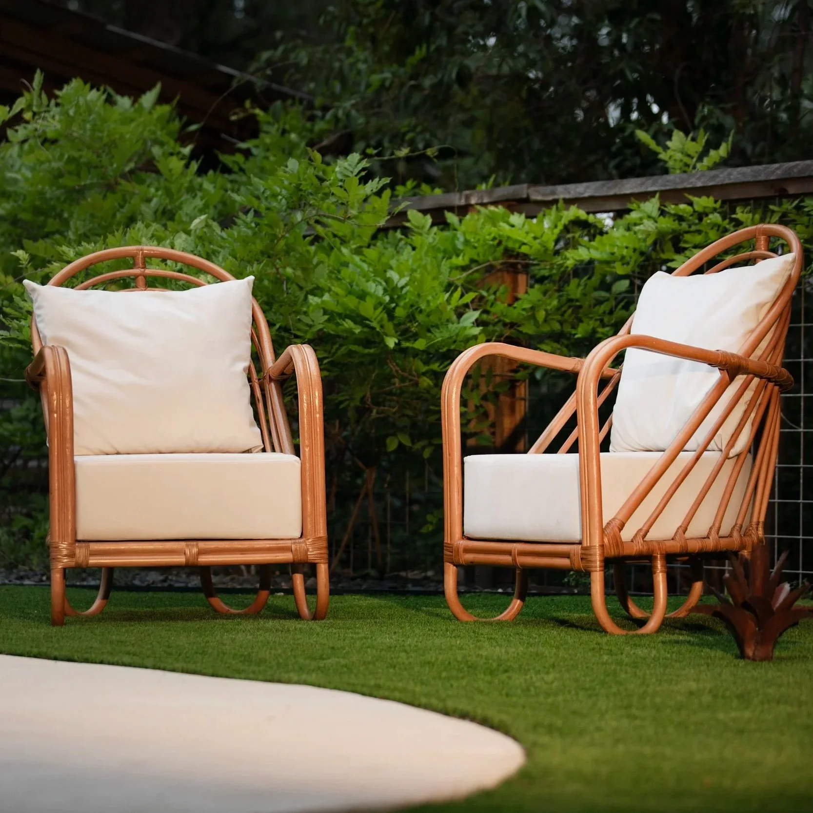 Sapa Rattan Chairs, Two wicker outdoor chairs with white cushions and pillows, set on a grass lawn with green bushes in the background.