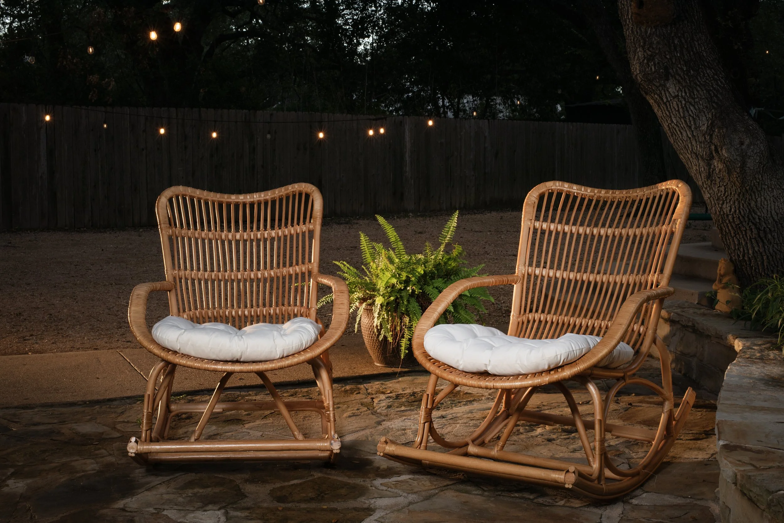 Two rattan rocking chairs with white cushions facing each other in a backyard yard at dusk, with string lights overhead, a wooden fence in the background, a potted fern between the chairs, and a large tree to the right.