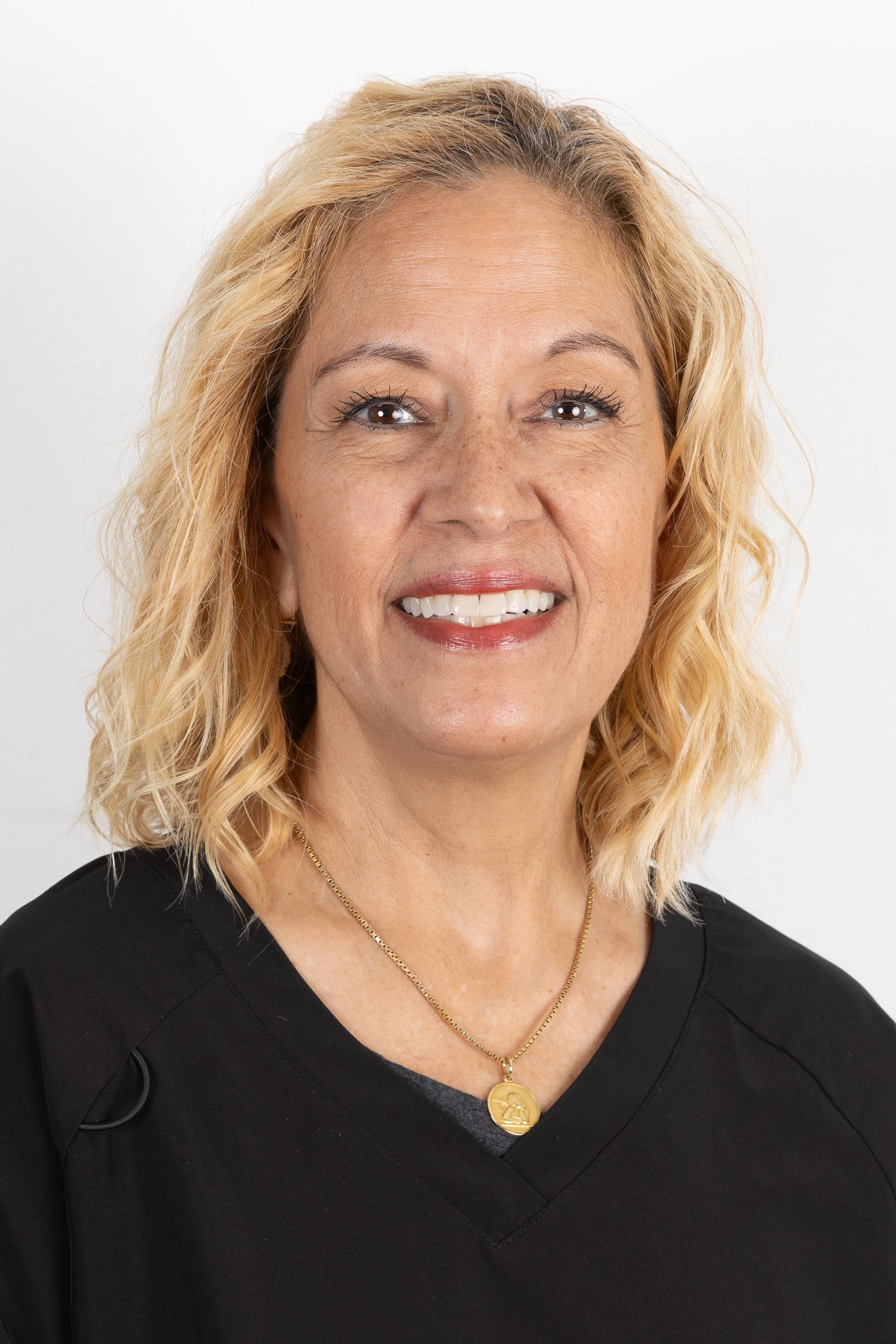 A middle-aged woman with shoulder-length blonde curly hair, wearing a black top and a gold necklace, smiling against a plain white background.