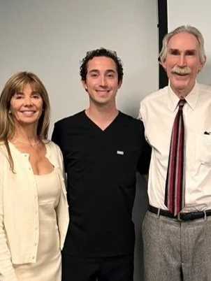 Three people standing close together indoors, smiling at the camera. The woman on the left has shoulder-length hair and is wearing a light-colored top and cardigan. The man in the middle has dark curly hair and is dressed in a black shirt. The man on the right has white hair and a mustache, wearing a white shirt with a striped tie and gray pants.