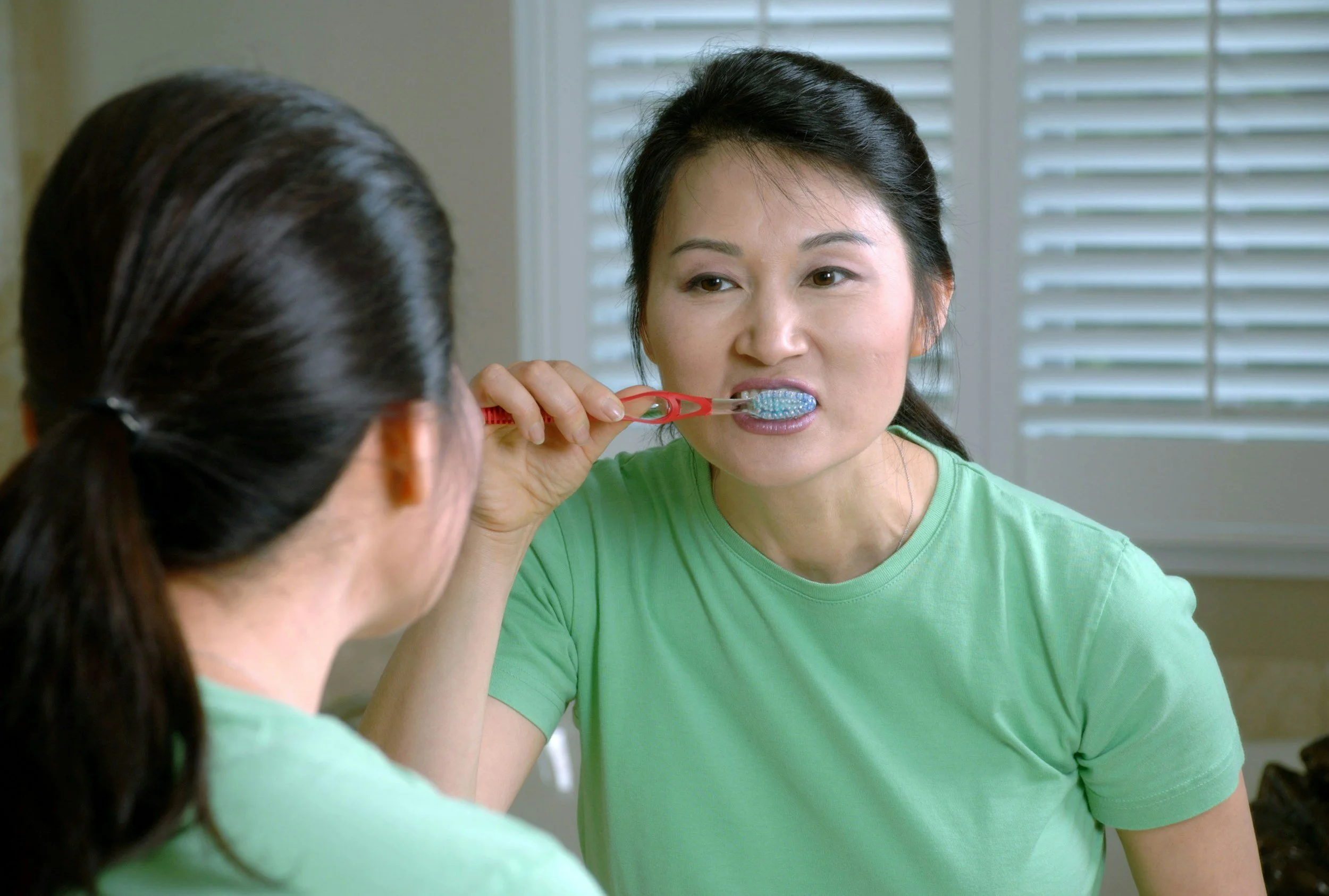 A woman brushing her teeth with another woman sitting in front of her, in a room with a window with blinds.