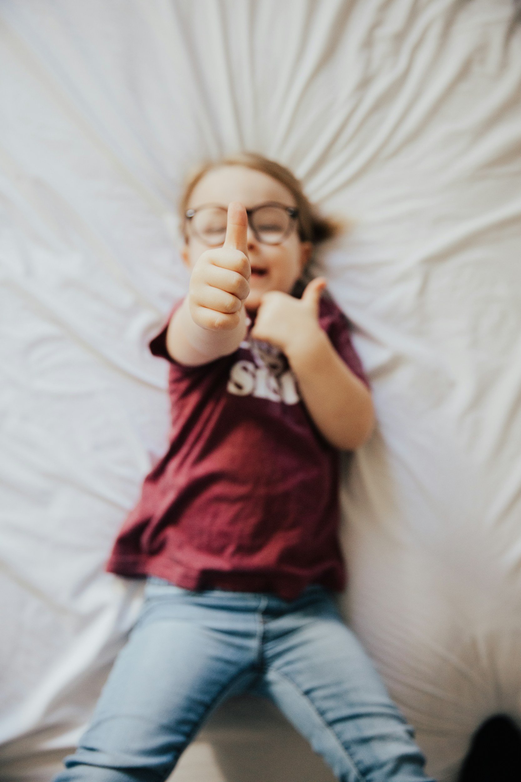 A young girl with glasses smiling and giving a thumbs-up while lying on a bed with white sheets.