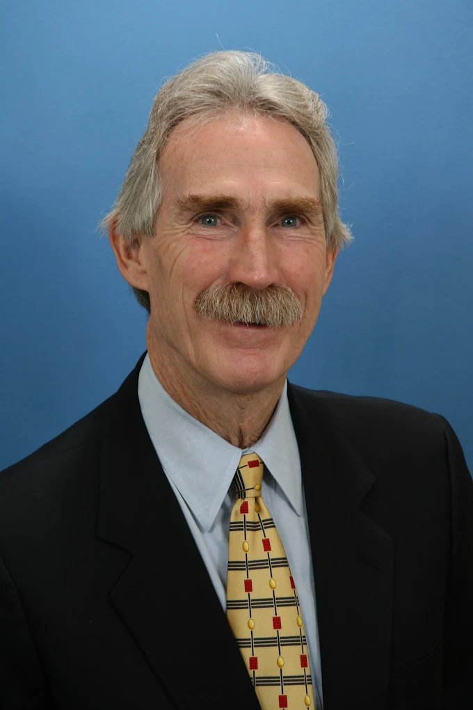 A man with gray hair and a mustache dressed in a suit and tie against a blue background.