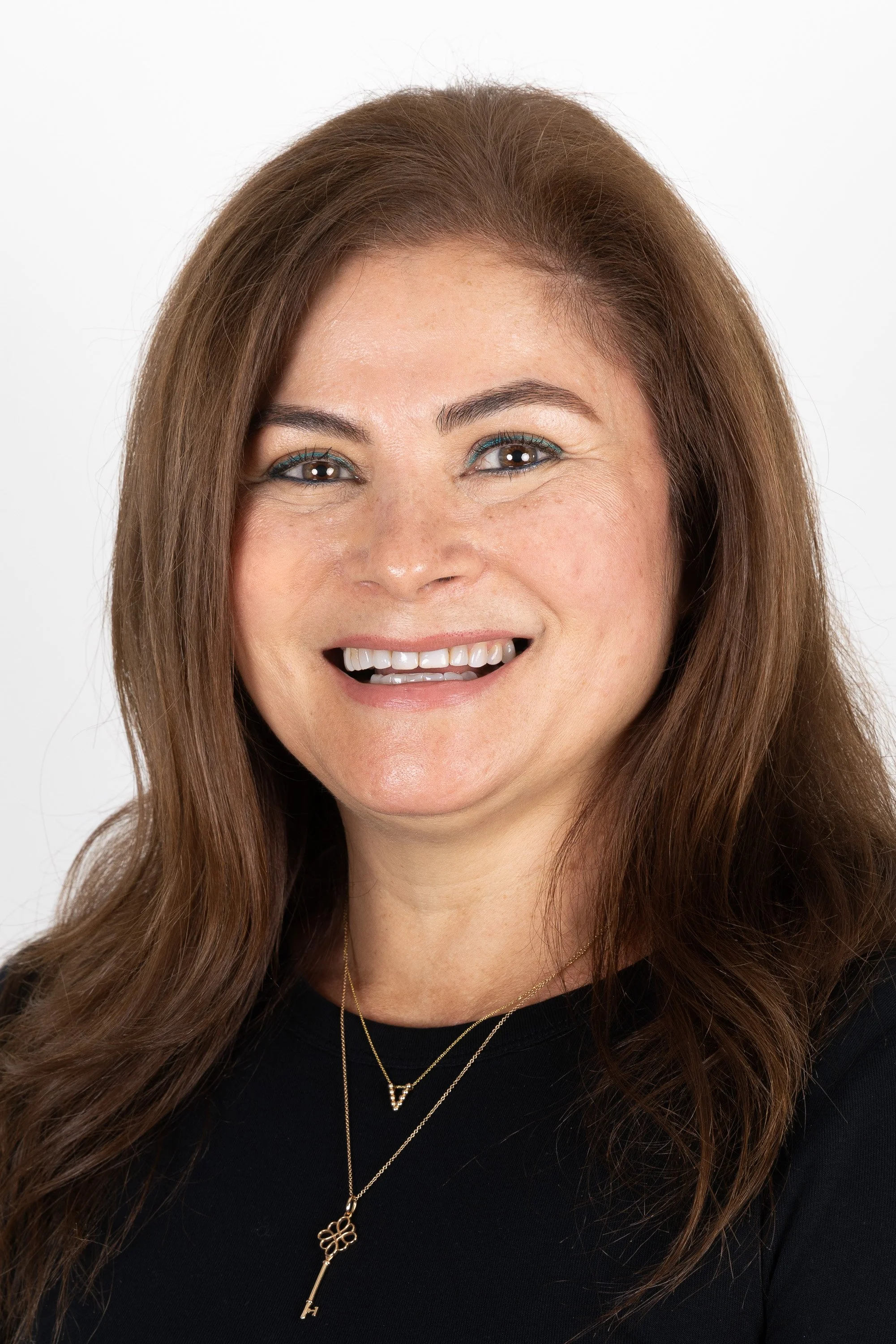 A woman with long, brown hair smiling and wearing a black top and layered necklaces.