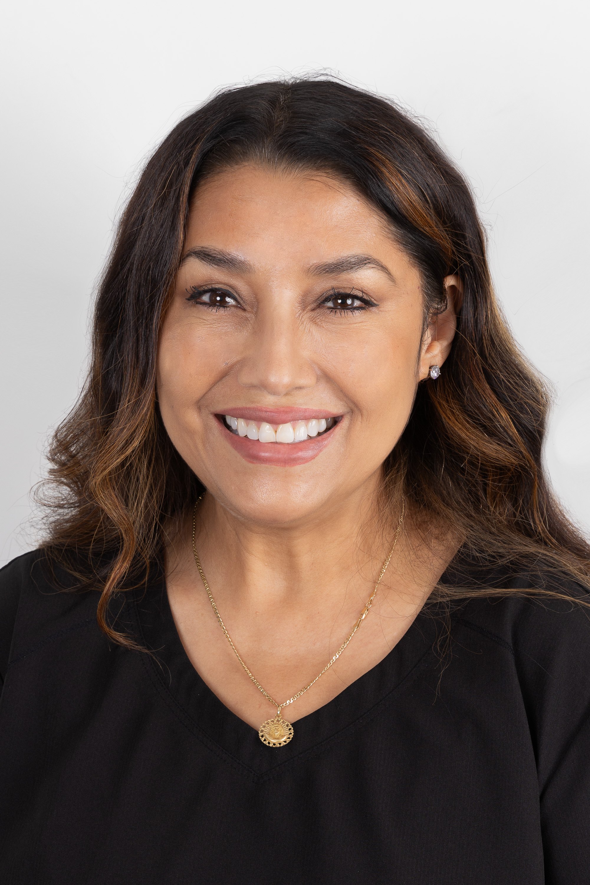 A smiling woman with shoulder-length brown hair, wearing a black top, earrings, and a gold necklace, against a plain white background.