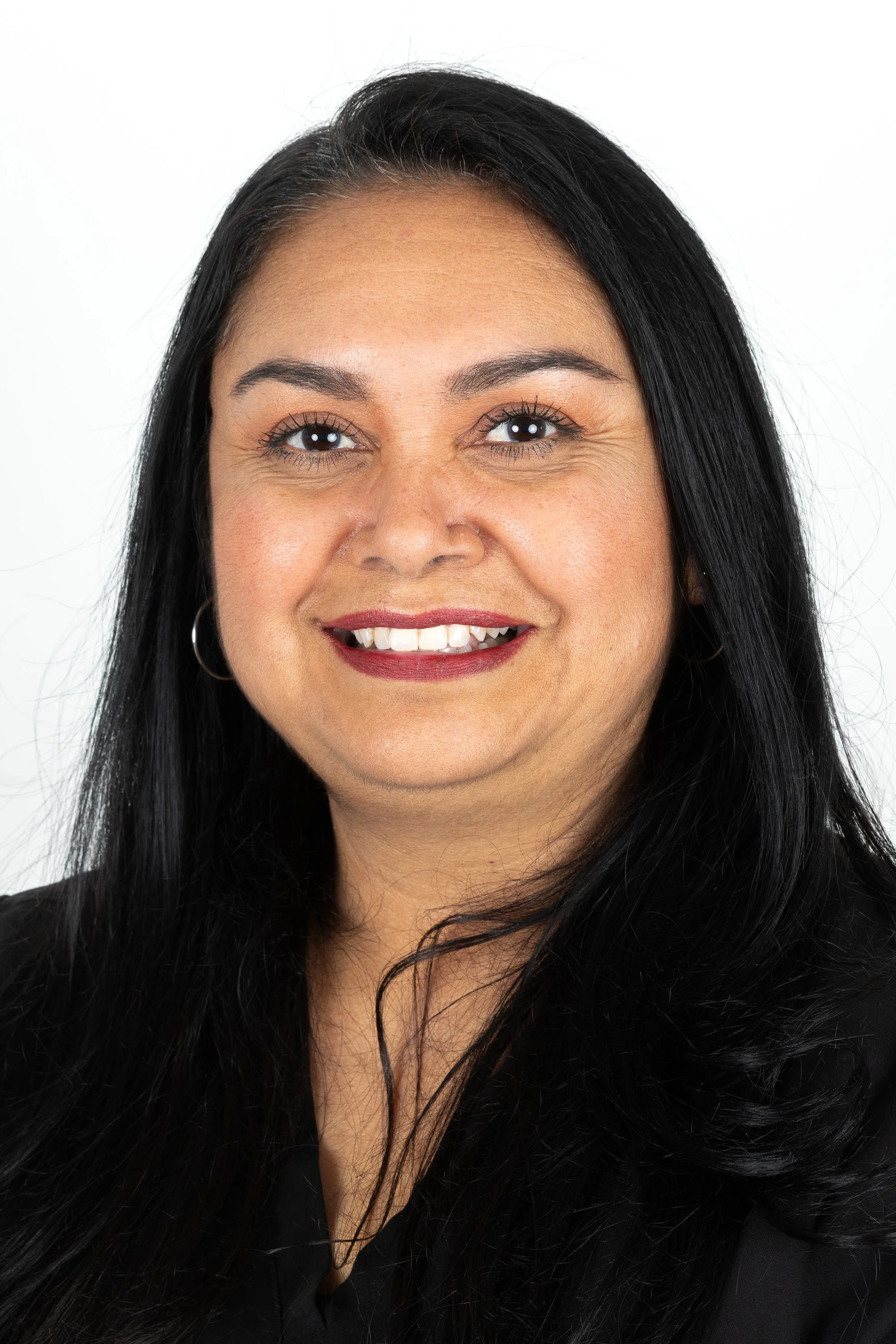 A woman with long black hair, wearing earrings, and a black top, smiling against a white background.