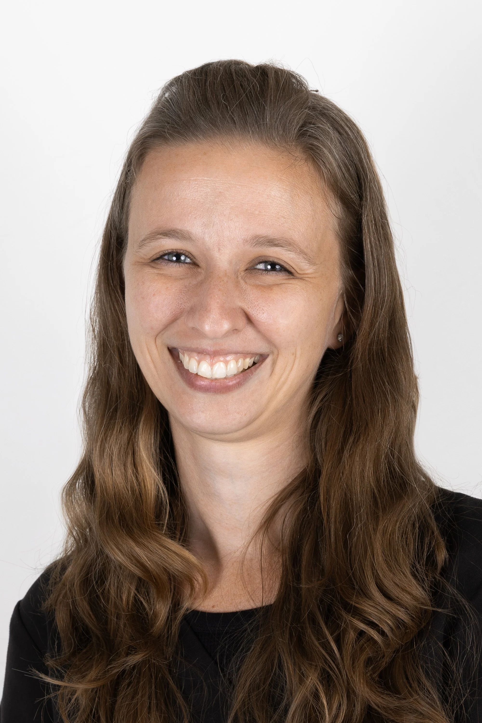 A smiling woman with long, wavy brown hair, wearing a black top, against a plain white background.