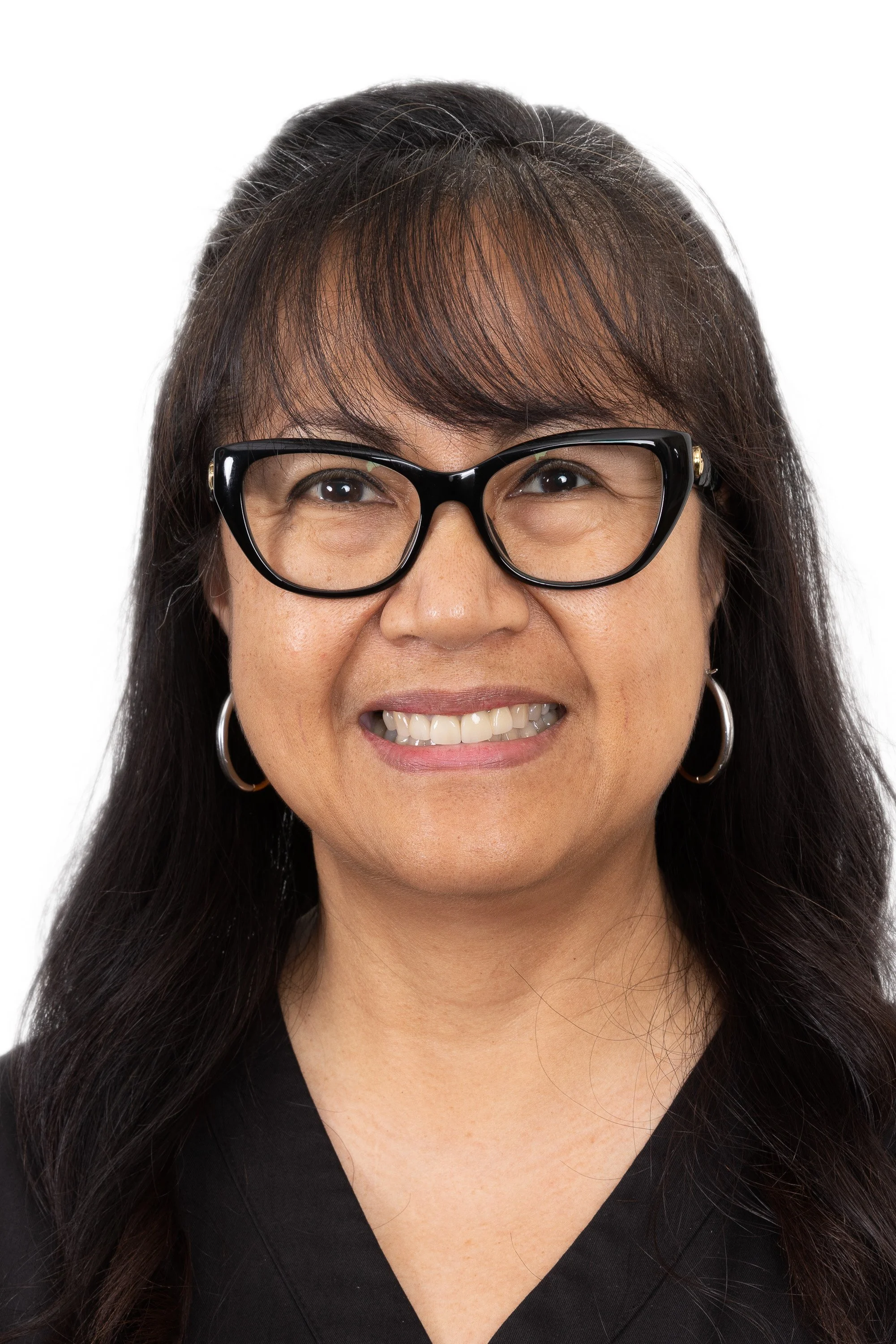 Portrait of a woman with long dark hair, wearing black glasses, hoop earrings, and a black top, smiling against a white background.