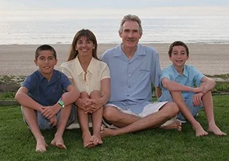 A family of four sitting on the grass by the beach, with ocean and sky in the background. The family includes a woman, a man, and two boys. They are all smiling and relaxed.