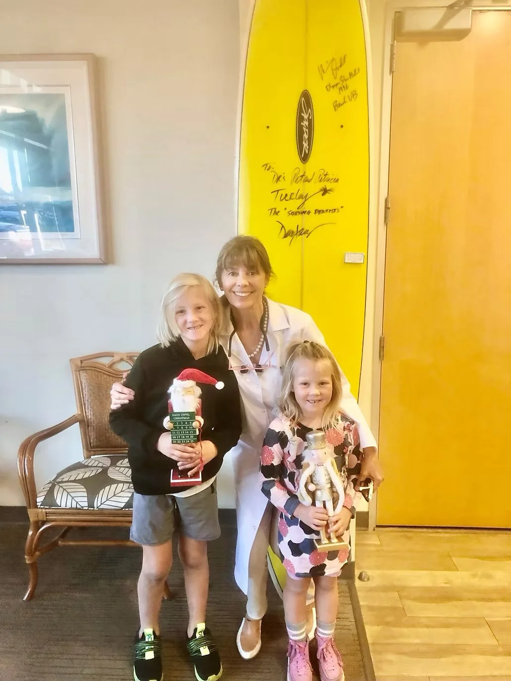 A woman with two young girls standing inside a room, holding holiday decorations, with a yellow surfboard signed by a famous person in the background.