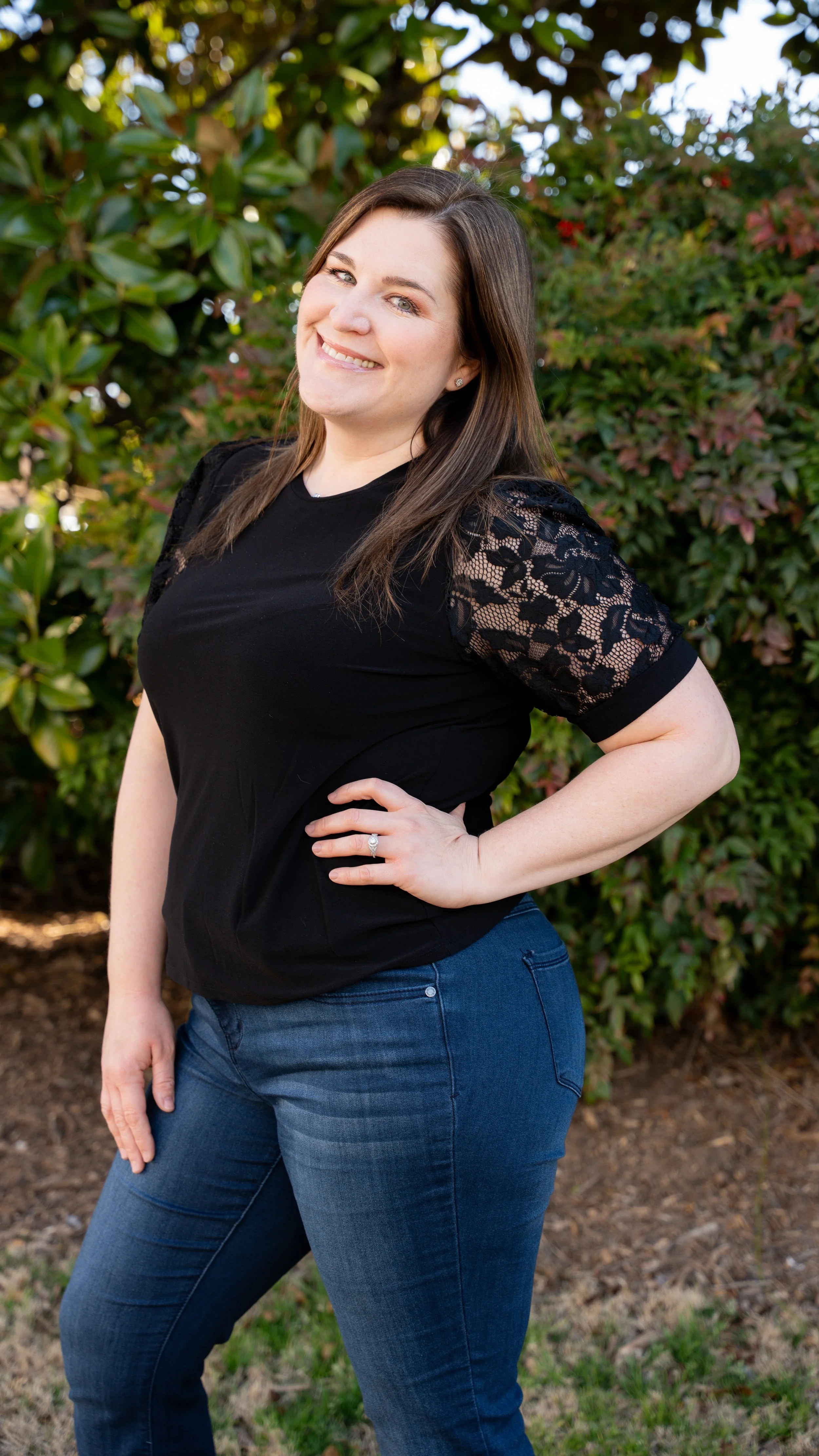 A woman standing outdoors near bushes, smiling, with her hand on her hip, wearing a black top with lace sleeves and blue jeans.