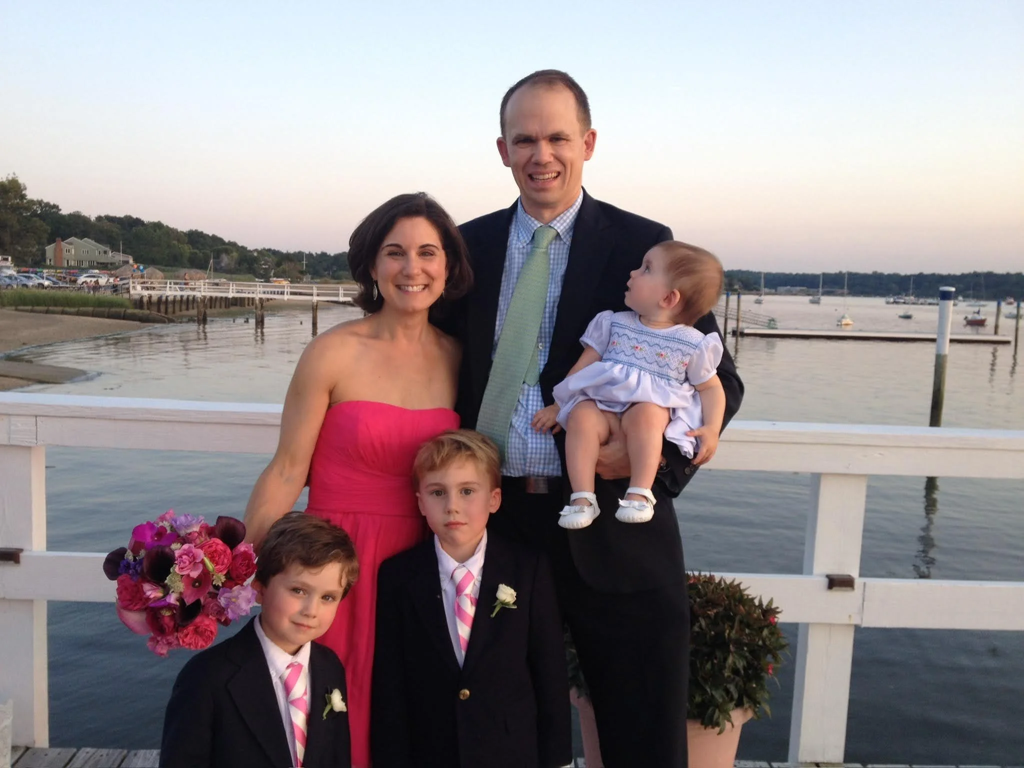Family of five stands on a pier near water at sunset, smiling, with boats and shoreline in background. The woman is in a pink strapless dress holding a bouquet, and the man is in a suit holding a toddler girl in a white dress. Two young boys in suits with pink-striped ties and white boutonnières stand in front.