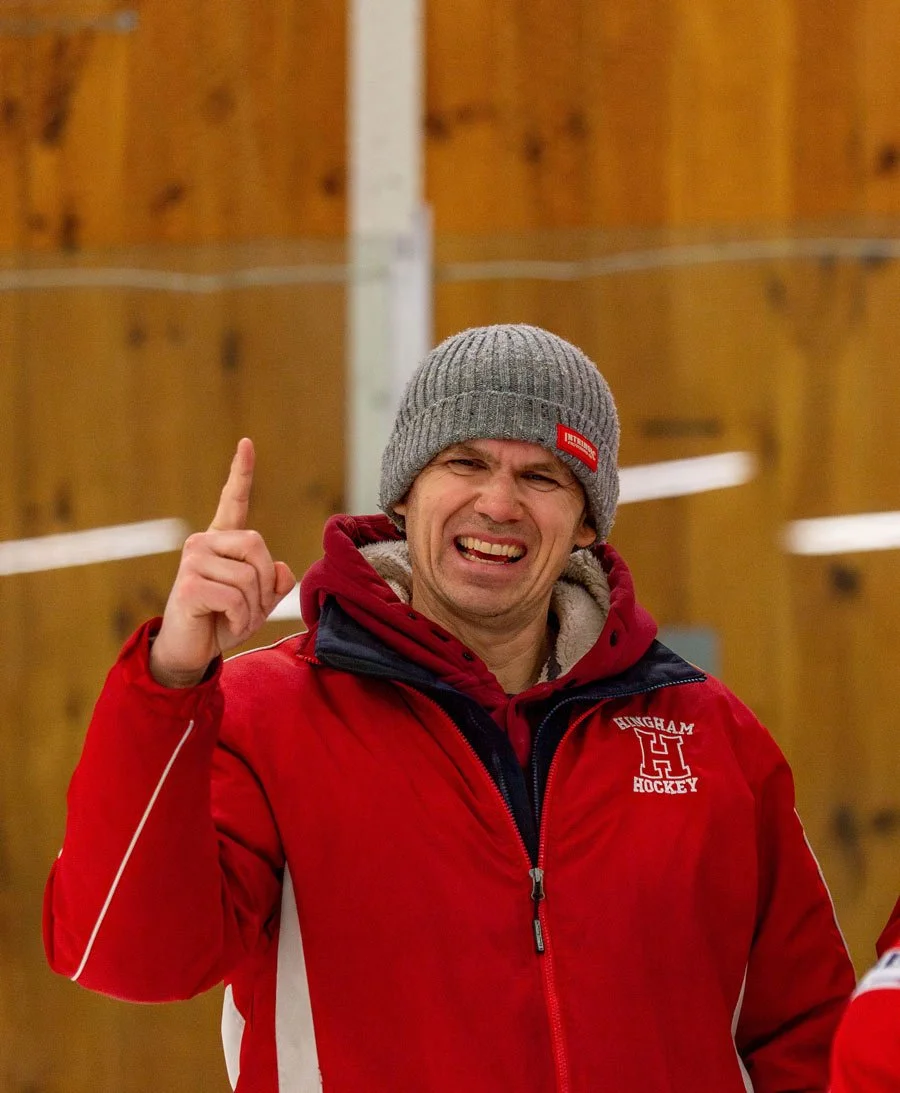 Rob Wittmann in red athletic jacket and grey knit beanie pointing upward and smiling in an indoor wooden ice rink setting.