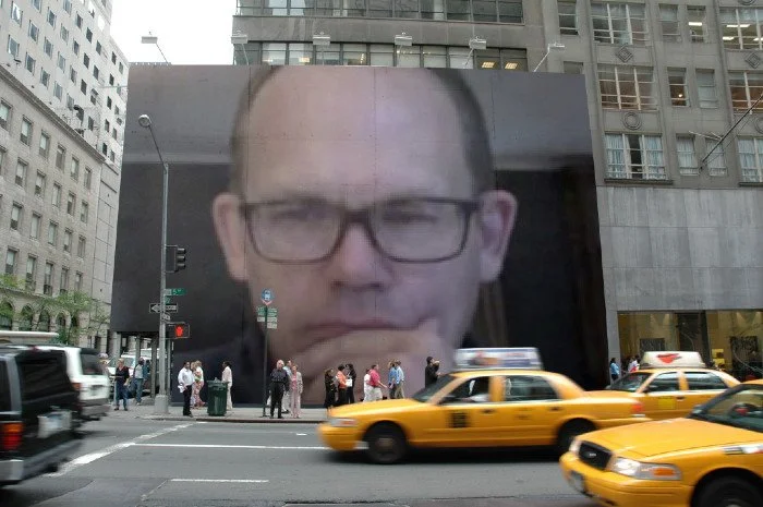 Large digital billboard displaying a close-up of a Rob Wittmann's face wearing glasses, with a serious expression, in an urban cityscape with taxis and people on the street.