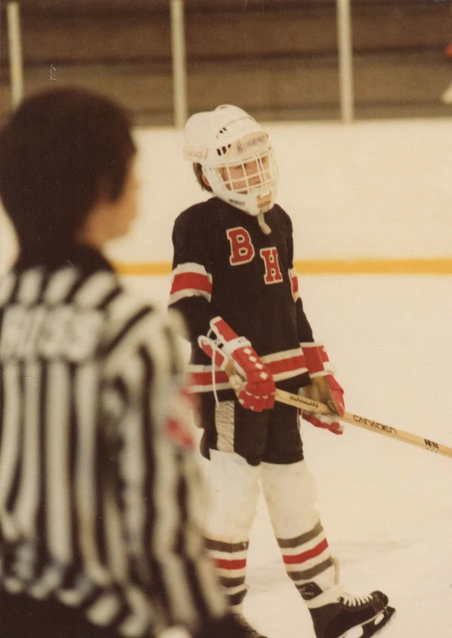 Rob Wittmann as a young hockey player in black and red uniform with the letters 'BH' on the front, wearing a white helmet and red gloves, standing on an ice rink holding a hockey stick, with a referee in black and white stripes in the foreground.